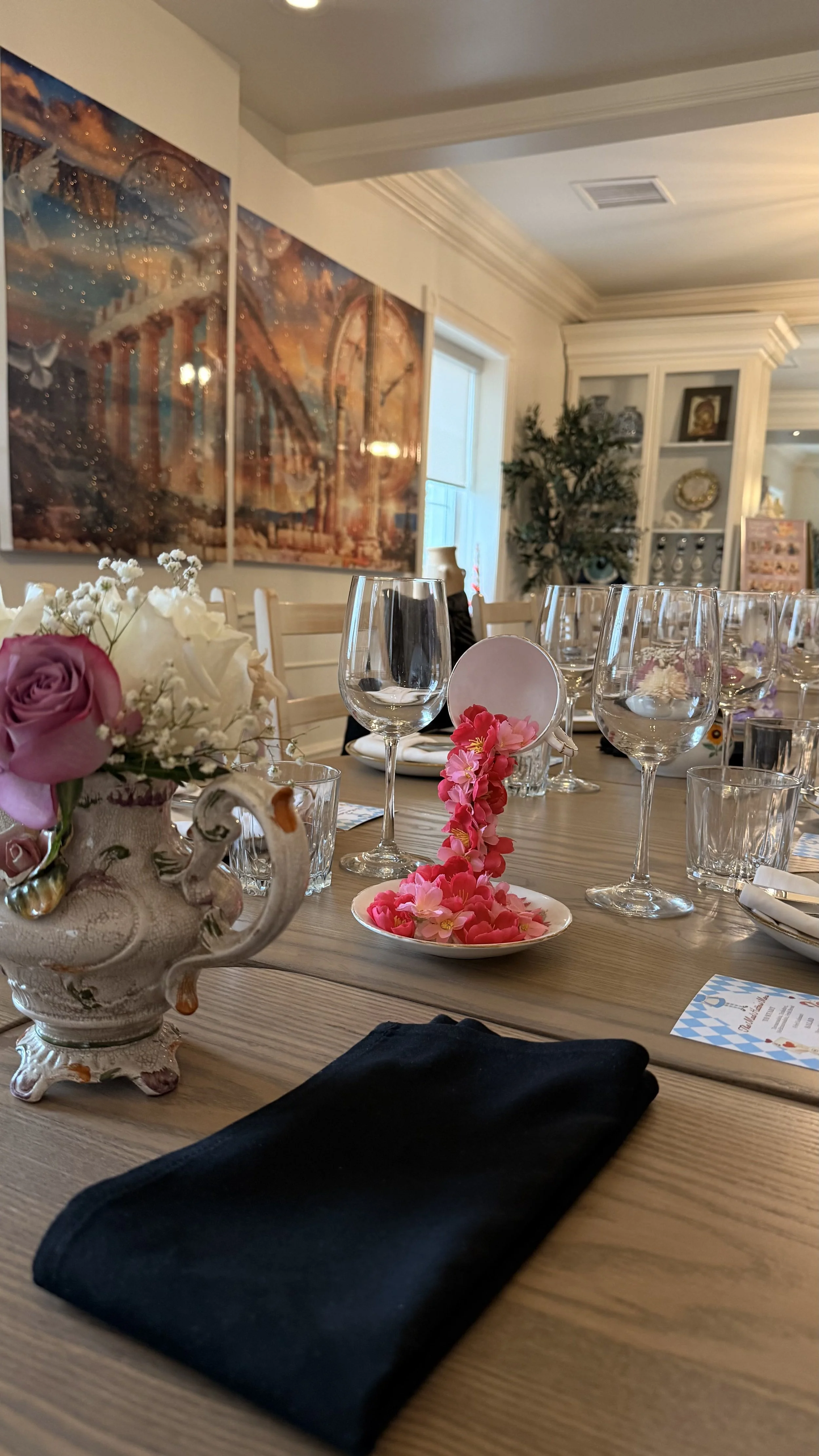 A dining table with a floral centerpiece, wine glasses, and pink flower decorations, set for a meal in a decorated room with artwork and shelves.