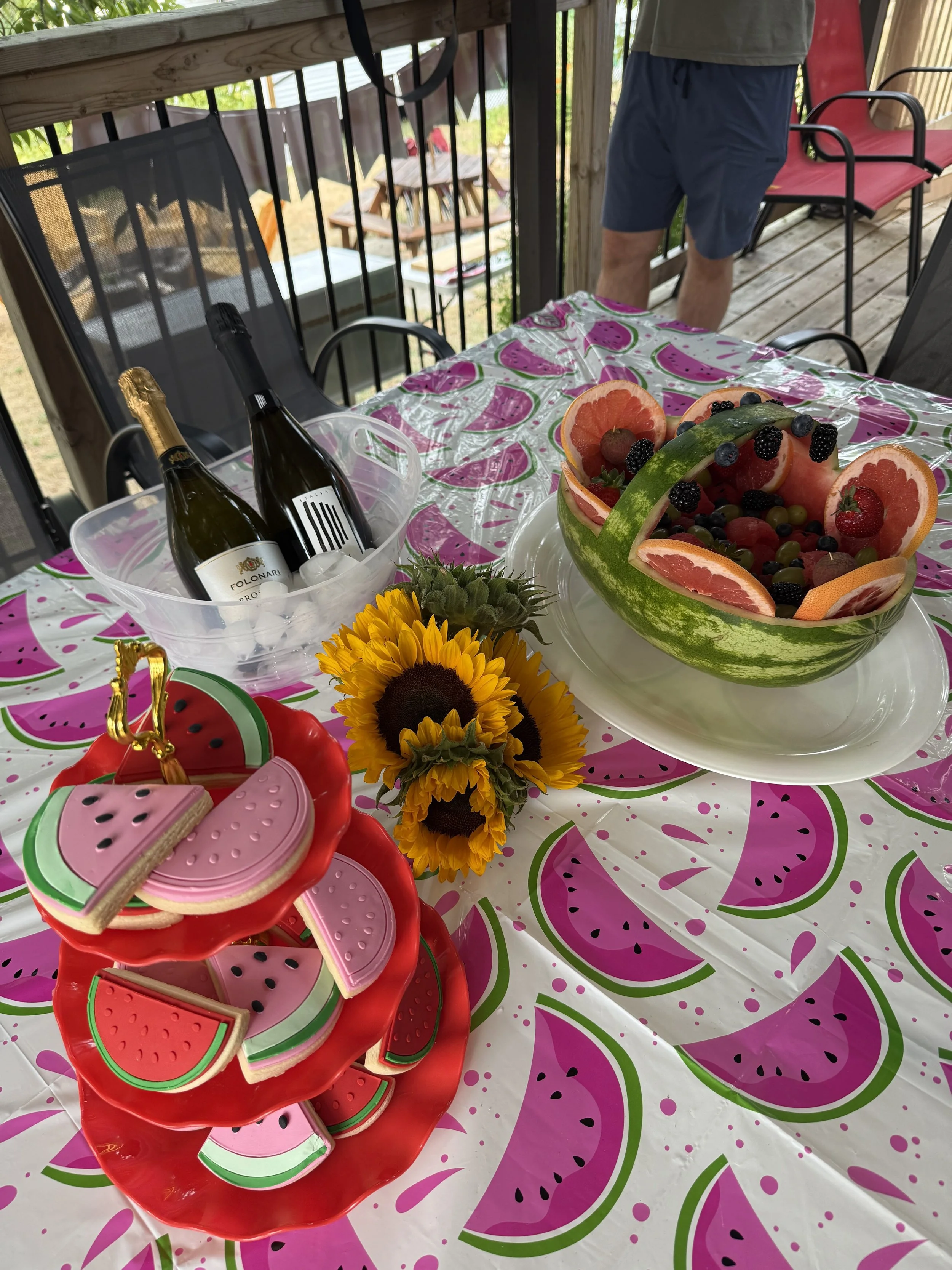 A table decorated with a watermelon-themed tablecloth, topped with a bowl of fresh fruit, a sunflower, and several watermelon-shaped cookies. In the background, a patio with chairs, a person standing, and bottles of sparkling cider or champagne in an