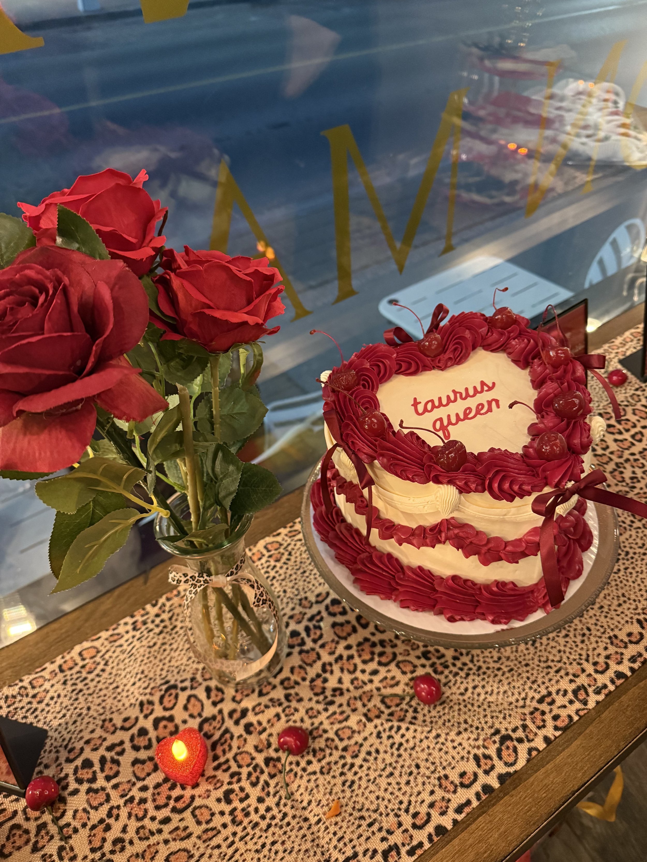 A centerpiece of six red roses in a glass vase, a decorated cake with red frosting, cherries, hearts, and a birthday message, placed on a leopard print tablecloth with small red cherries and a red rose-shaped candle nearby.