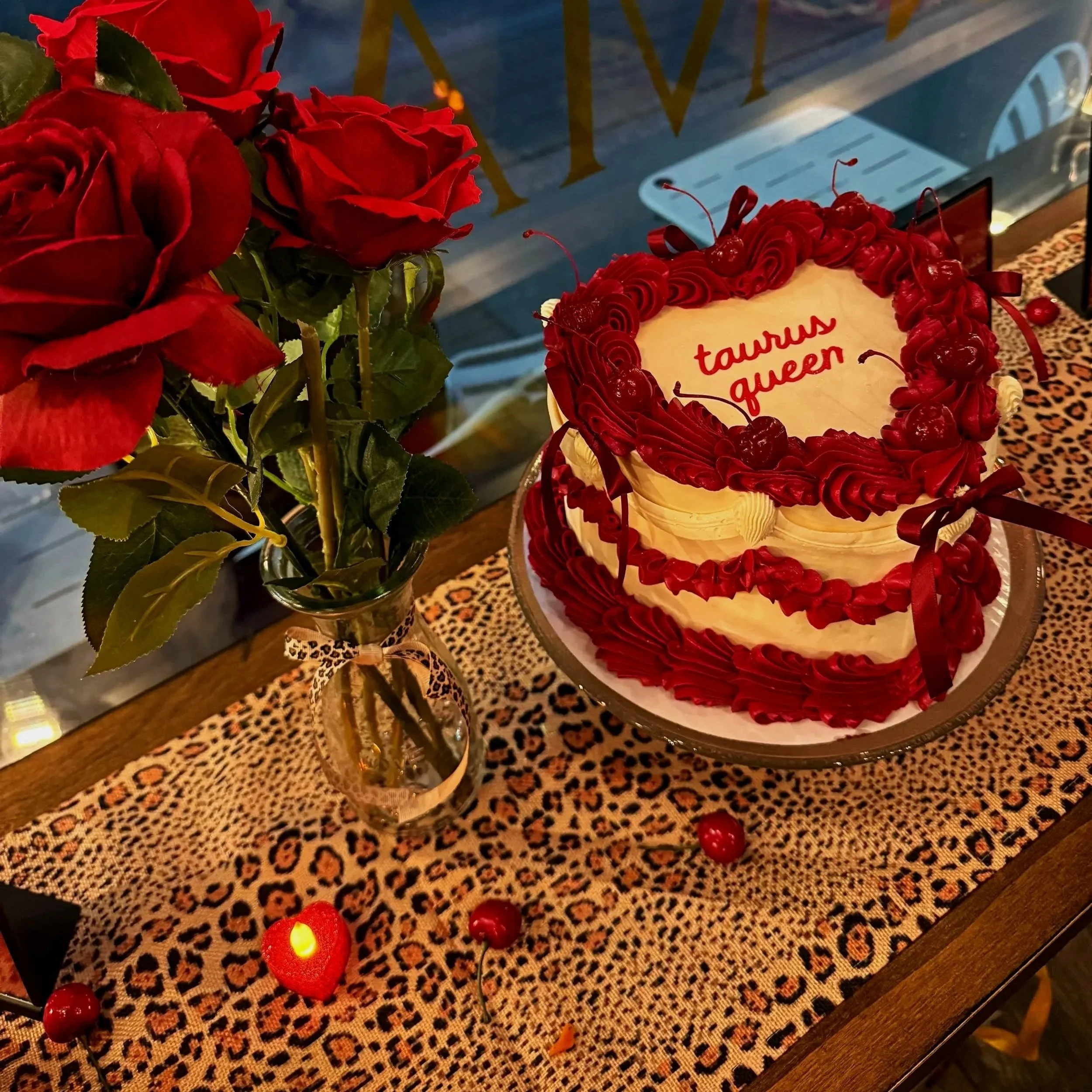 Red roses in a vase and a heart-shaped cake with red frosting and cherries, with the words "tawwes aem" written on it, sitting on a leopard print tablecloth.