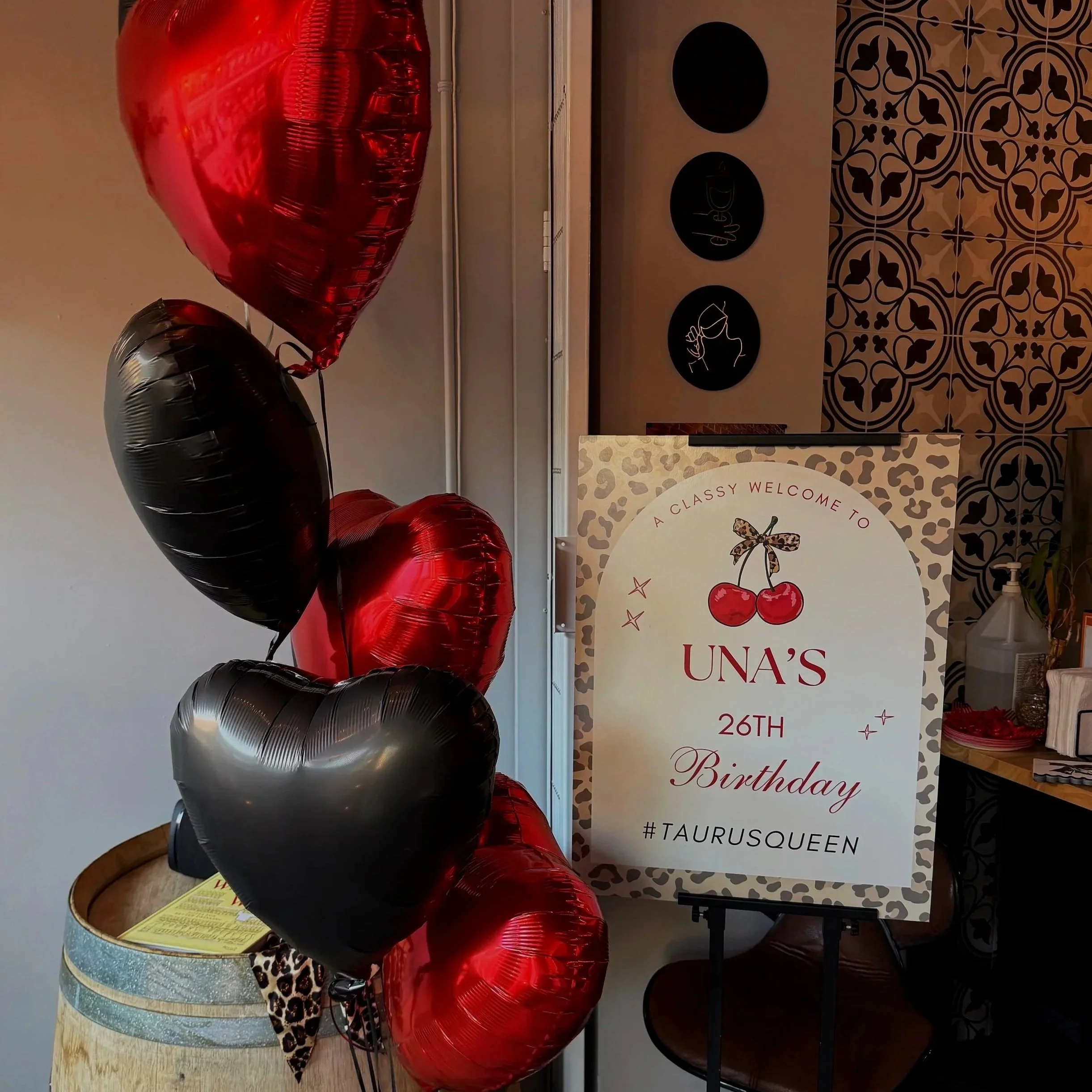 Balancing act with multiple balloons in red, black, and silver, set against a wellness-themed backdrop, including a banner with cherries and the words "A classy welcome toUna's 26th birthday," and decorative wall patterns.