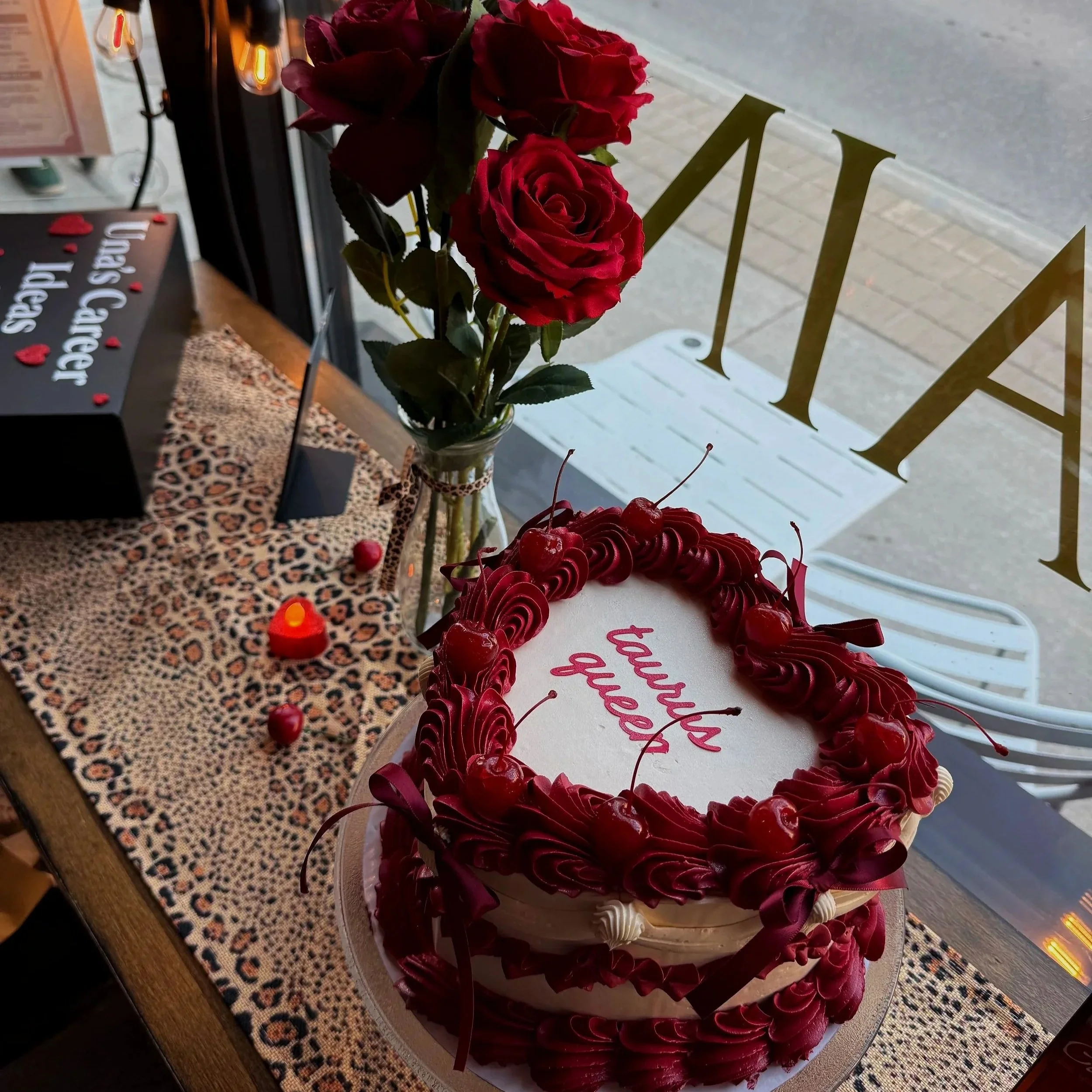A round cake decorated with red roses, cherries, and red ribbon, with a white base, and a message that says 'Happy Valentine' on top, displayed in a bakery or café window.