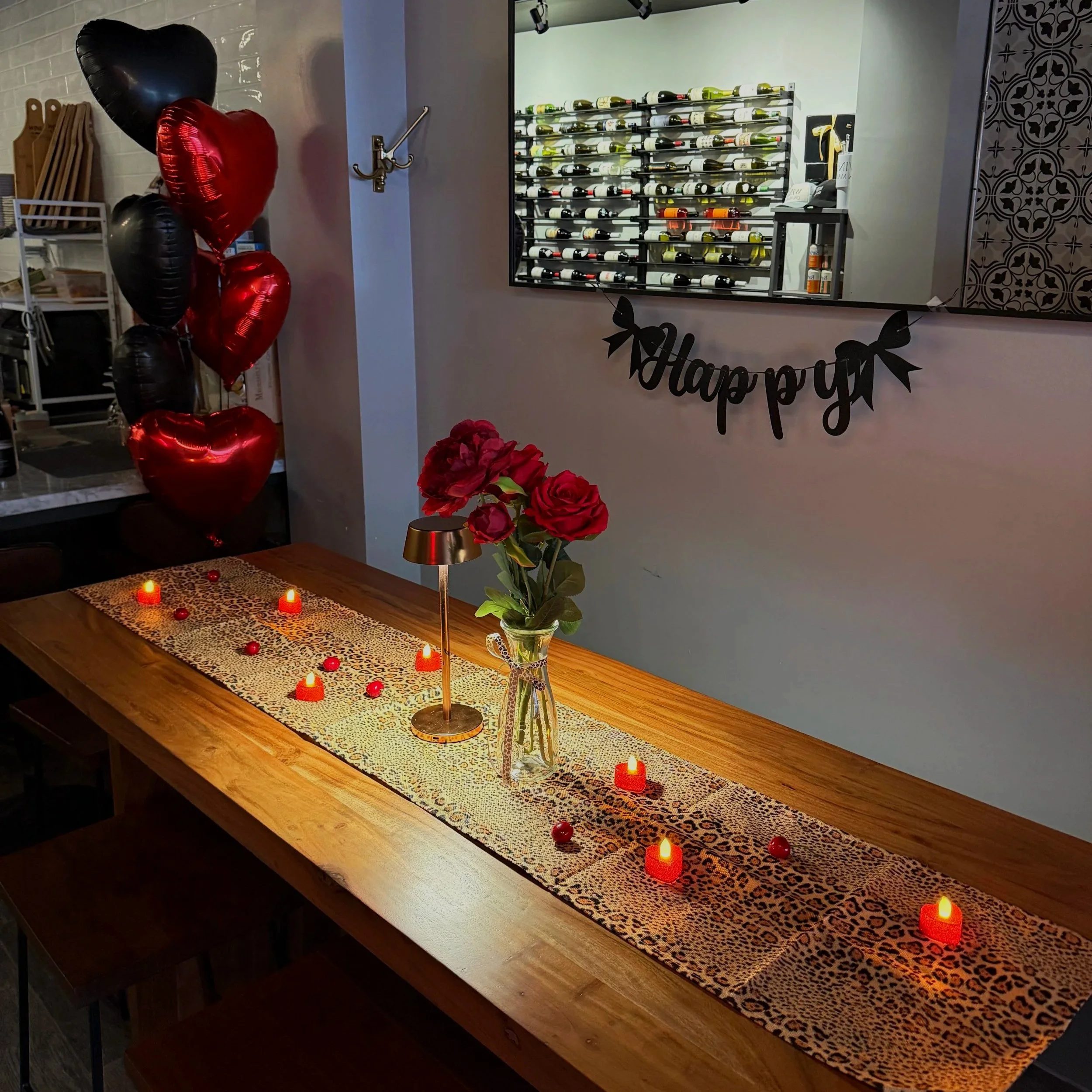 A table decorated for Valentine's Day with red candles and a red and leopard print table runner. In the background, black and red heart-shaped balloons and a large mirror showing a wine rack with bottles. A sign hanging reads 'Happy' with an arrow.