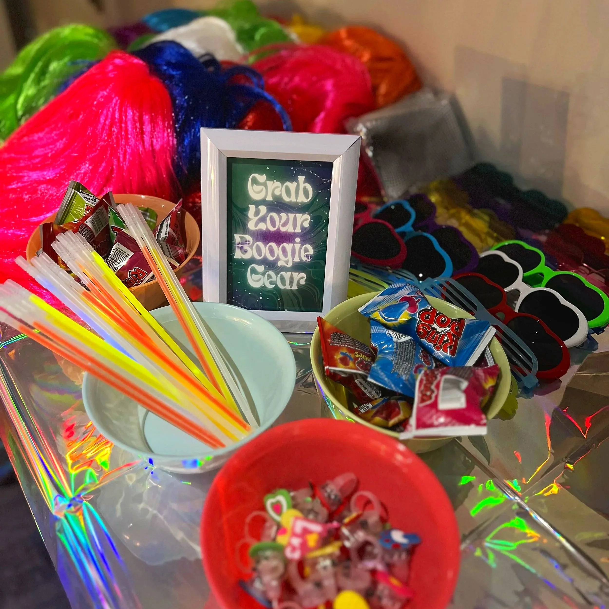 Colorful loom bands in a clear container with a sign that says 'Grab Your Boogie Gear', surrounded by various candy, straws, and toys.