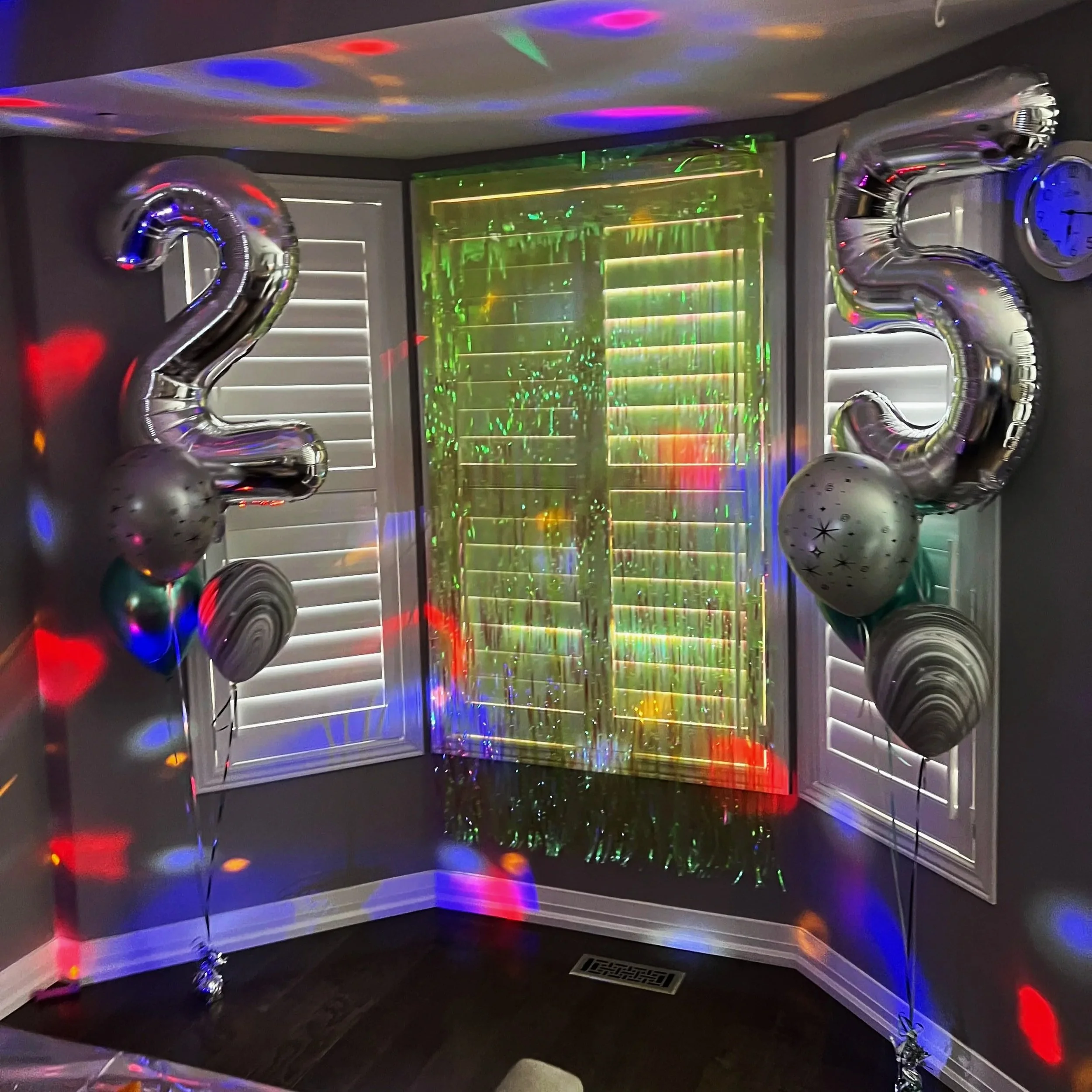 Silver balloons shaped as the number 23 are displayed in a room with reflective surfaces and colorful lights projected on the walls and ceiling, creating a festive atmosphere.