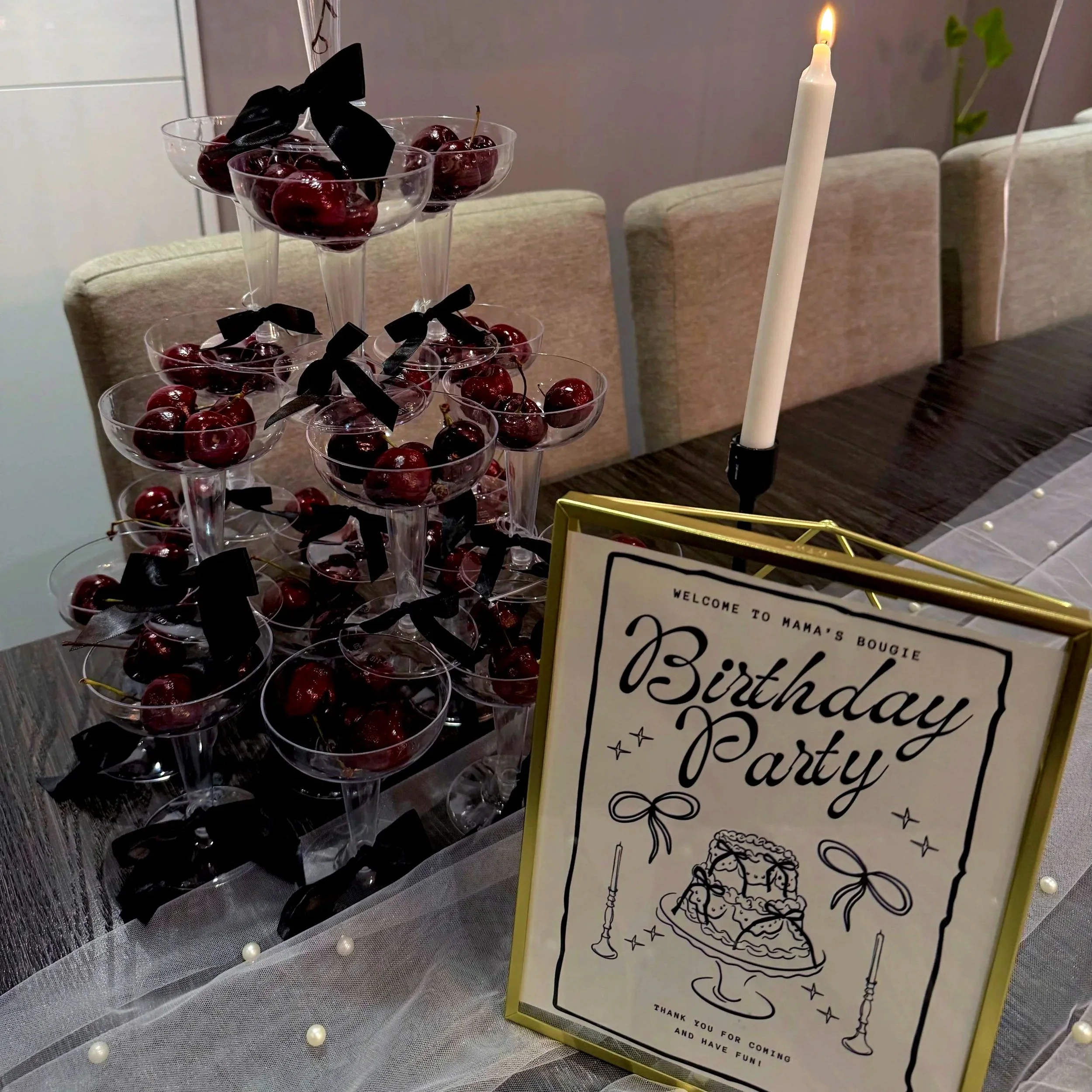 A multi-tiered stand with glasses filled with cherries and black ribbon bows, likely for a birthday celebration. In the background, a lit birthday candle and a decorative sign on a table with tulle and pearl accents are visible, indicating a festive 
