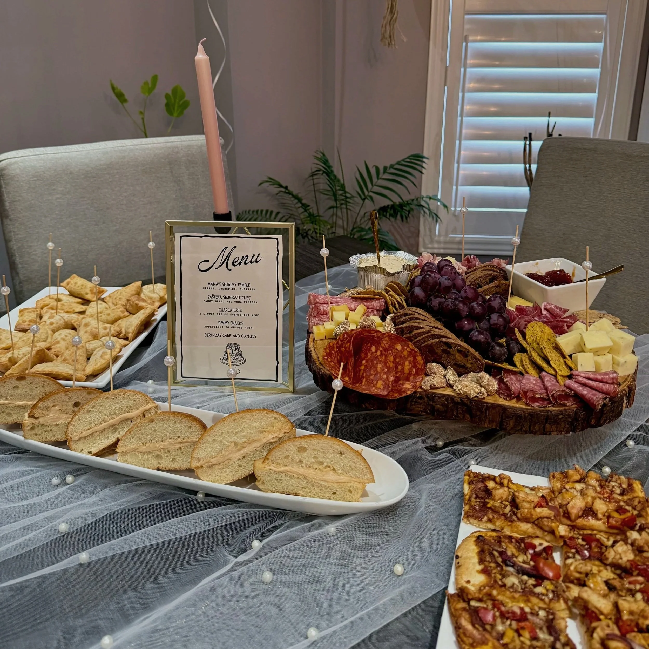 A table with assorted snacks including crackers, cheese, grapes, cured meats, and a dessert bar. In the background, there are chairs, a window with blinds, and a candle.