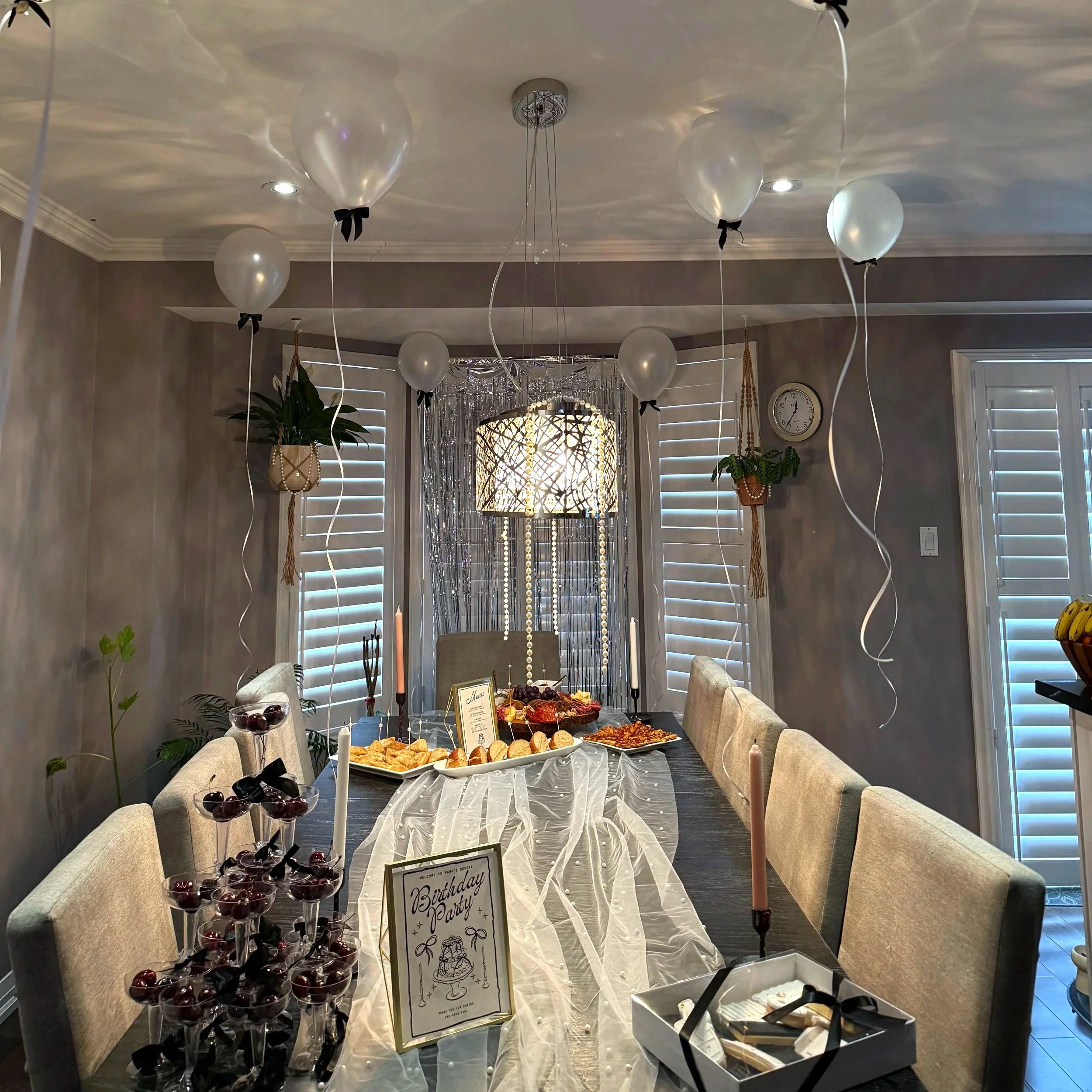 Indoor birthday party setup with a long table covered with a white tablecloth, surrounded by beige chairs, featuring various desserts, a sign that reads 'Birthday Party,' and balloons attached to the ceiling.