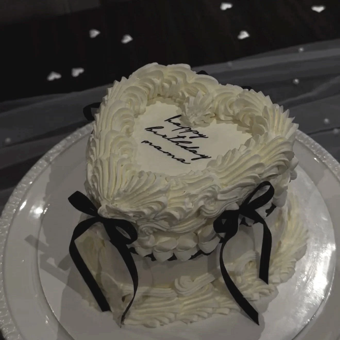A round white cake with piped frosting decorations, black ribbon bows, and a message on top that reads 'happy birthday mama,' on a white plate with a silver border.