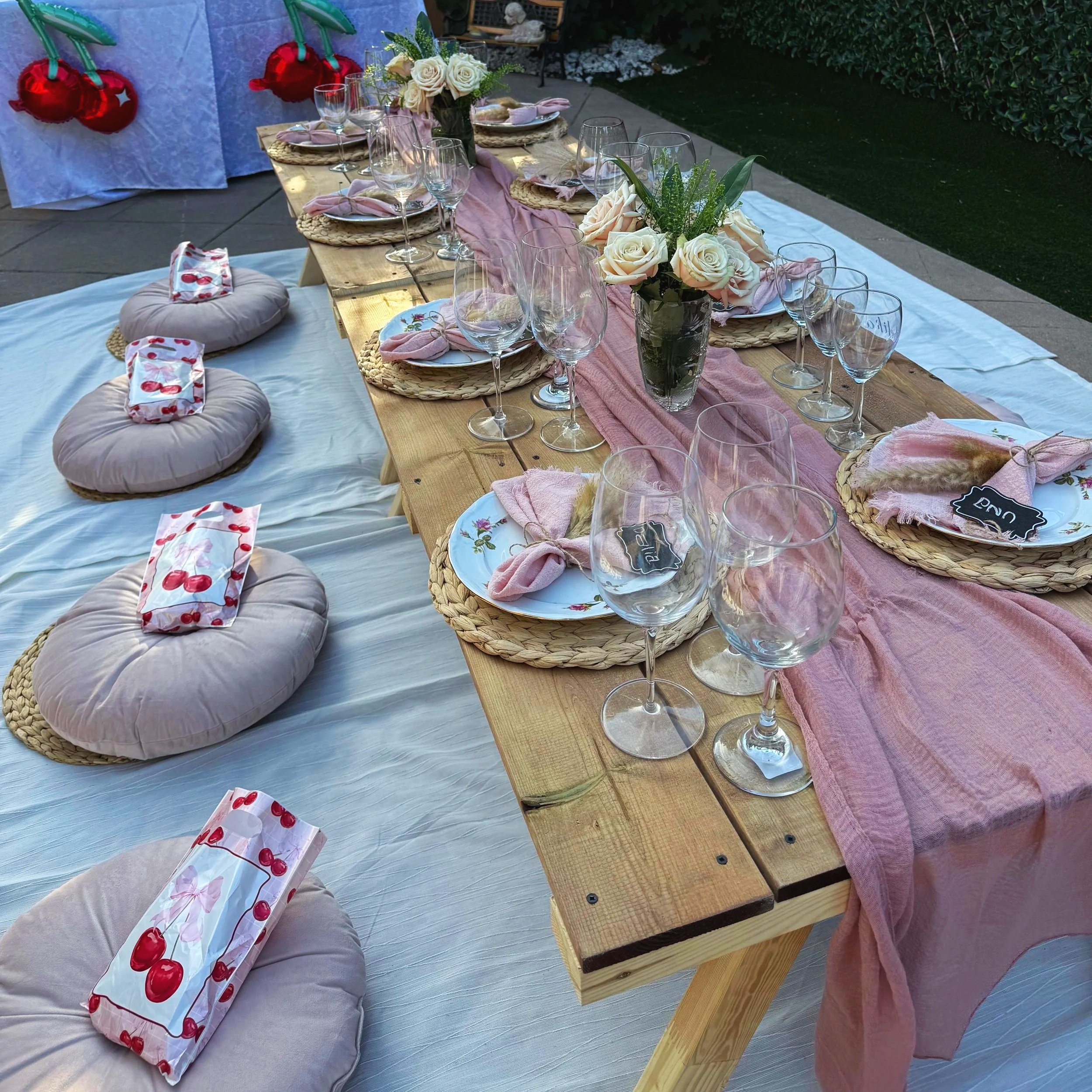 Outdoor dining setup with a wooden table decorated with pink table runner, floral arrangements, and glassware, on a patio with a white cloth underneath. There are floor cushions with cherry-patterned pillows and an area with balloons in the backgroun