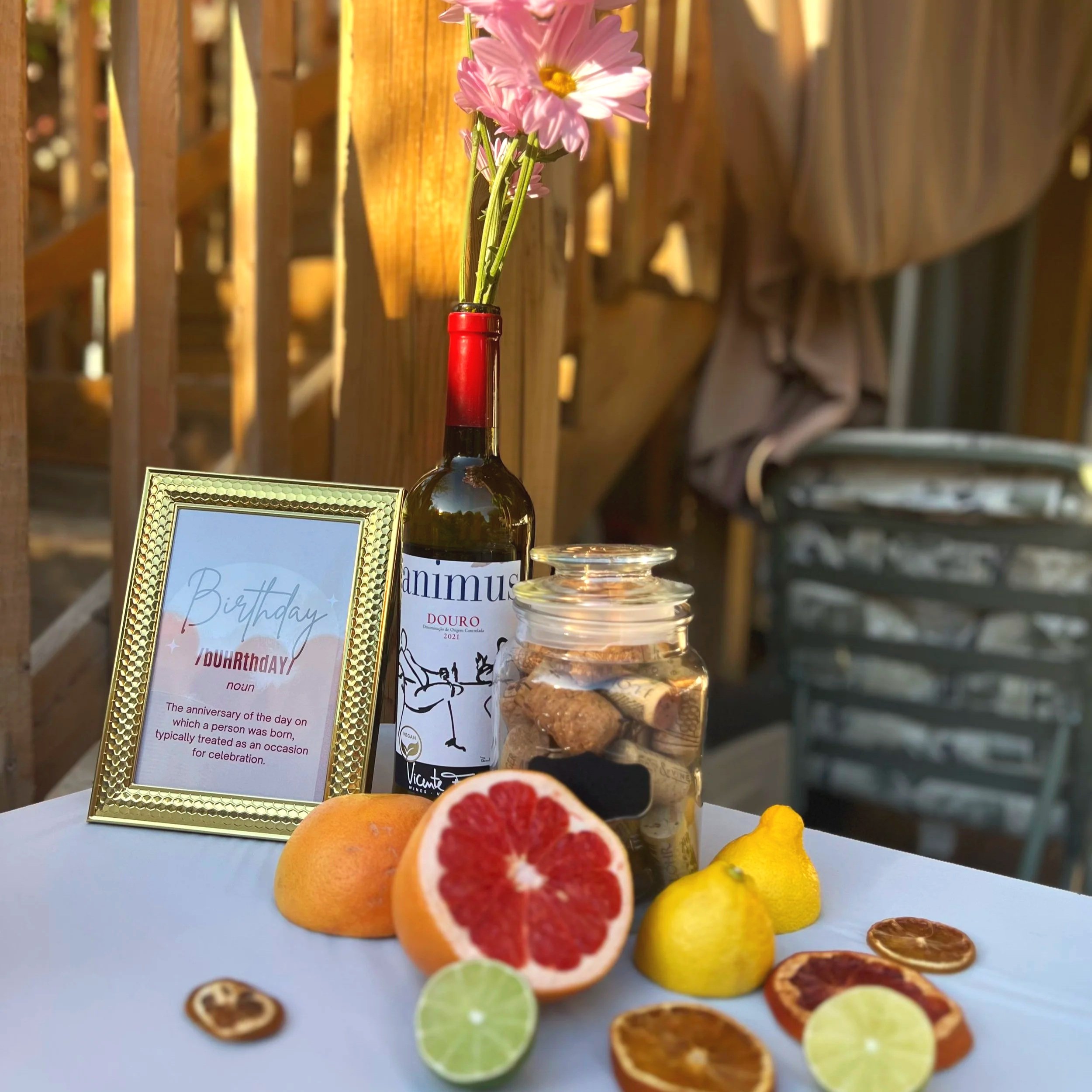 A wine bottle with pink and yellow artificial flowers in the neck, next to a framed birthday message and a variety of toy fruits, against a wooden fence background.