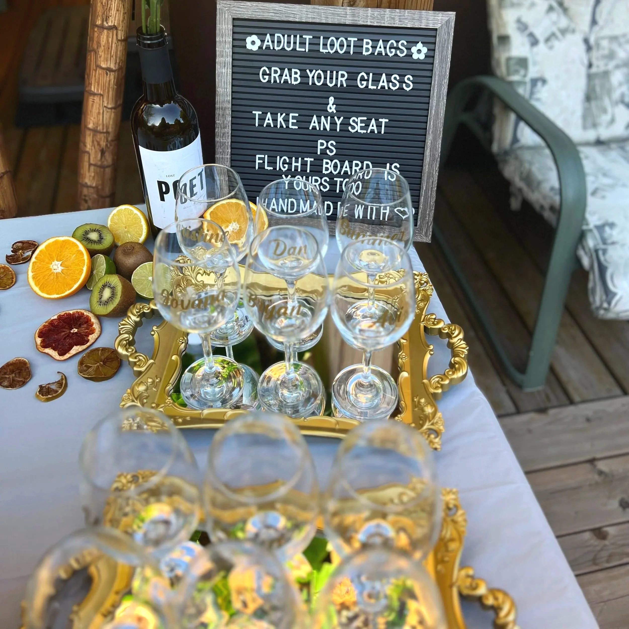 A signboard with instructions to grab an adult loot bag and a display of sliced fruits including oranges, kiwis, and dried citrus slices, with a bottle of wine and glasses in the foreground.