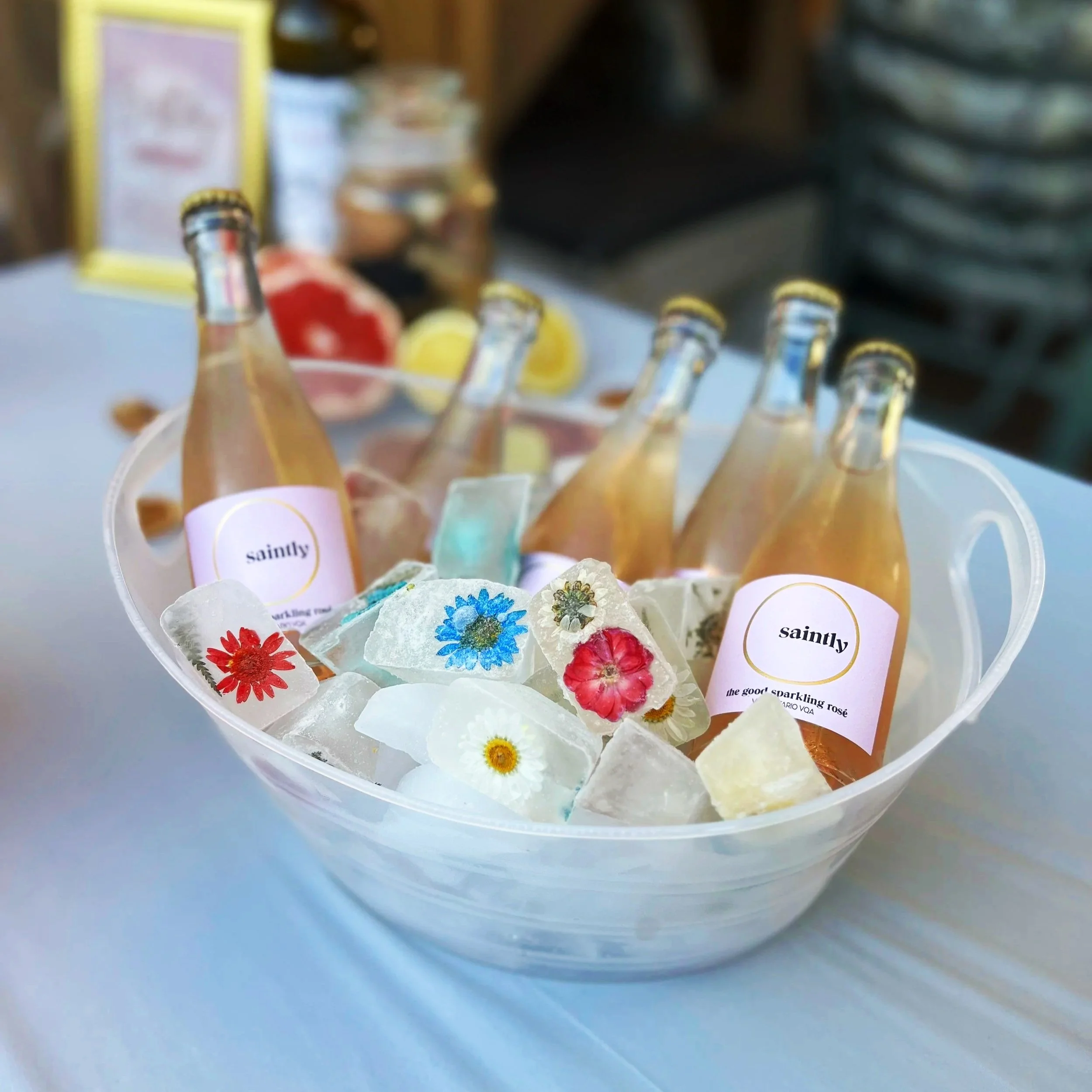 Close-up of small clear bottles with yellow caps on a white surface, with colorful flowers and labels that say 'saintly' and 'the good sparkling rose' in the foreground.