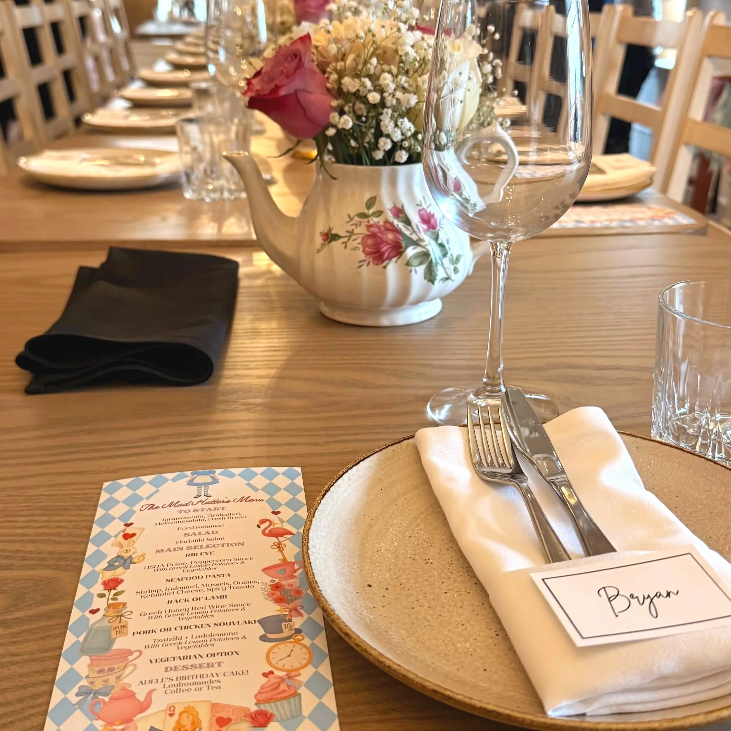 A dining table setup with a floral centerpiece featuring pink and white flowers in a ceramic teapot, elegant glassware including a wine glass, a menu card, and a plate with a napkin and cutlery. The background shows wooden chairs and a cozy restauran
