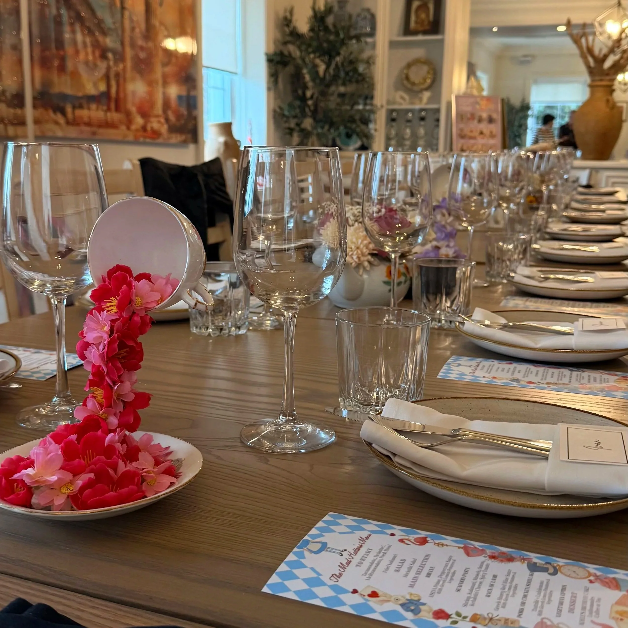 A table set with empty wine glasses, a plate of pink and red flowers, and a colorful menu, inside a decorated room with paintings, shelves, and large vases.