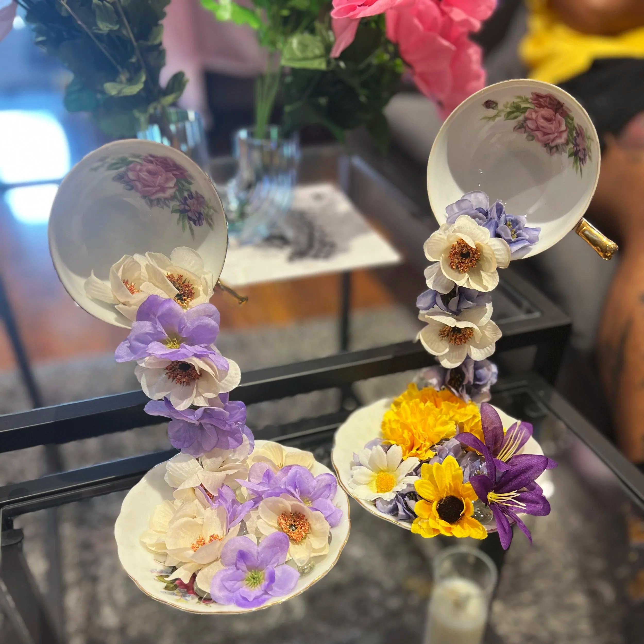 Close-up of a flower arrangement with small porcelain cups decorated with pink roses, purple, yellow, and white flowers, and green foliage on a black metal stand.