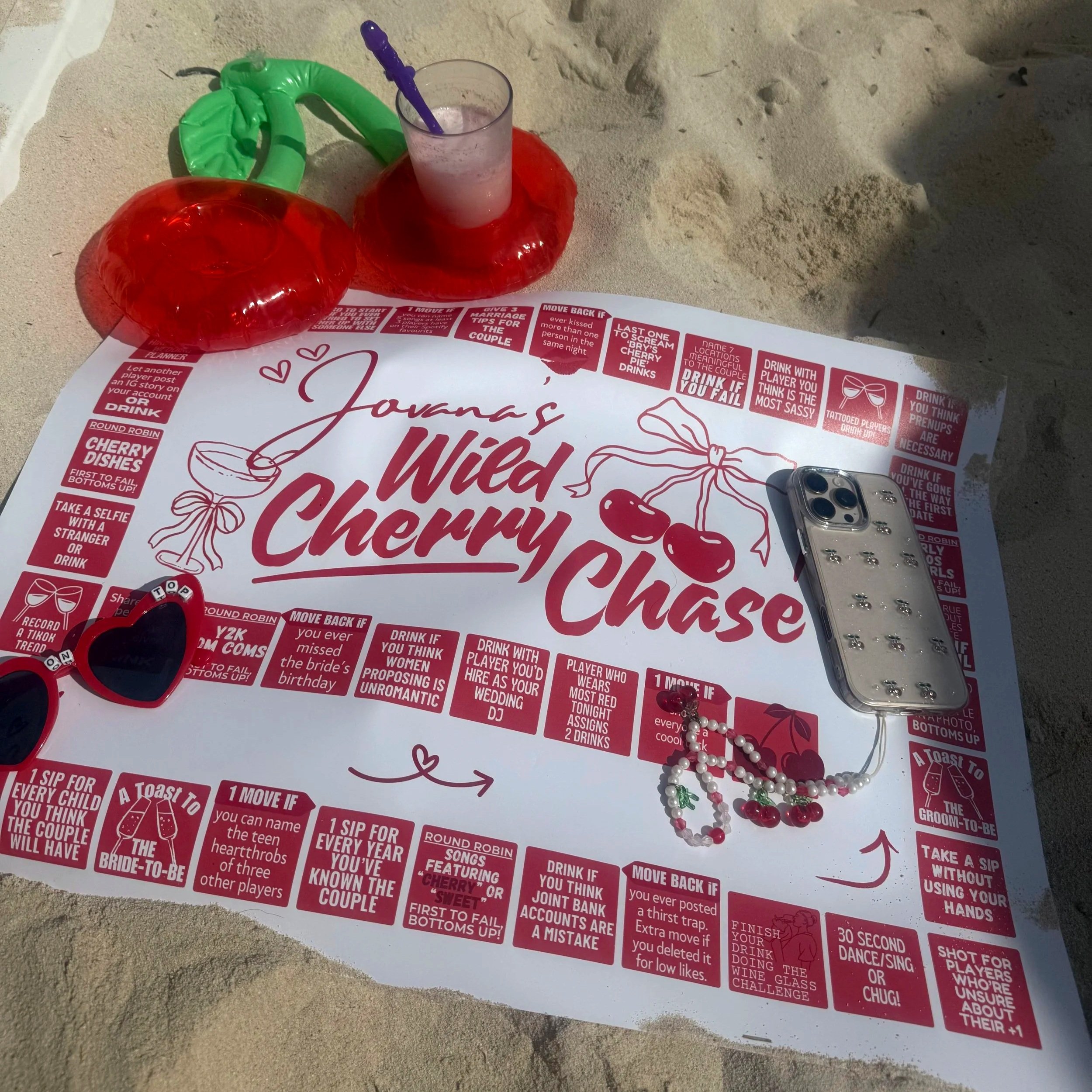 A beach scene with a printed game sheet titled 'How Well Do You Know the Couple' spread on sand, surrounded by a green inflatable ring, red heart-shaped sunglasses, a pearl necklace with a cherry charm, a silver phone case, and a glass with a purple 