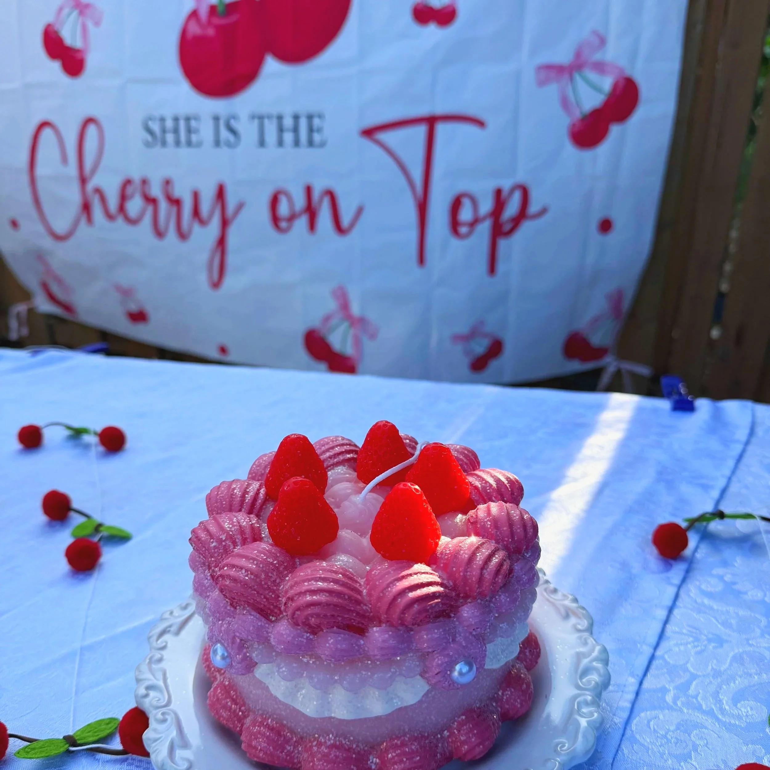 A cake decorated with pink frosting and red heart-shaped candies, placed on a white decorative platter. The background has a card with red and pink cherry illustrations and red text, partially visible.