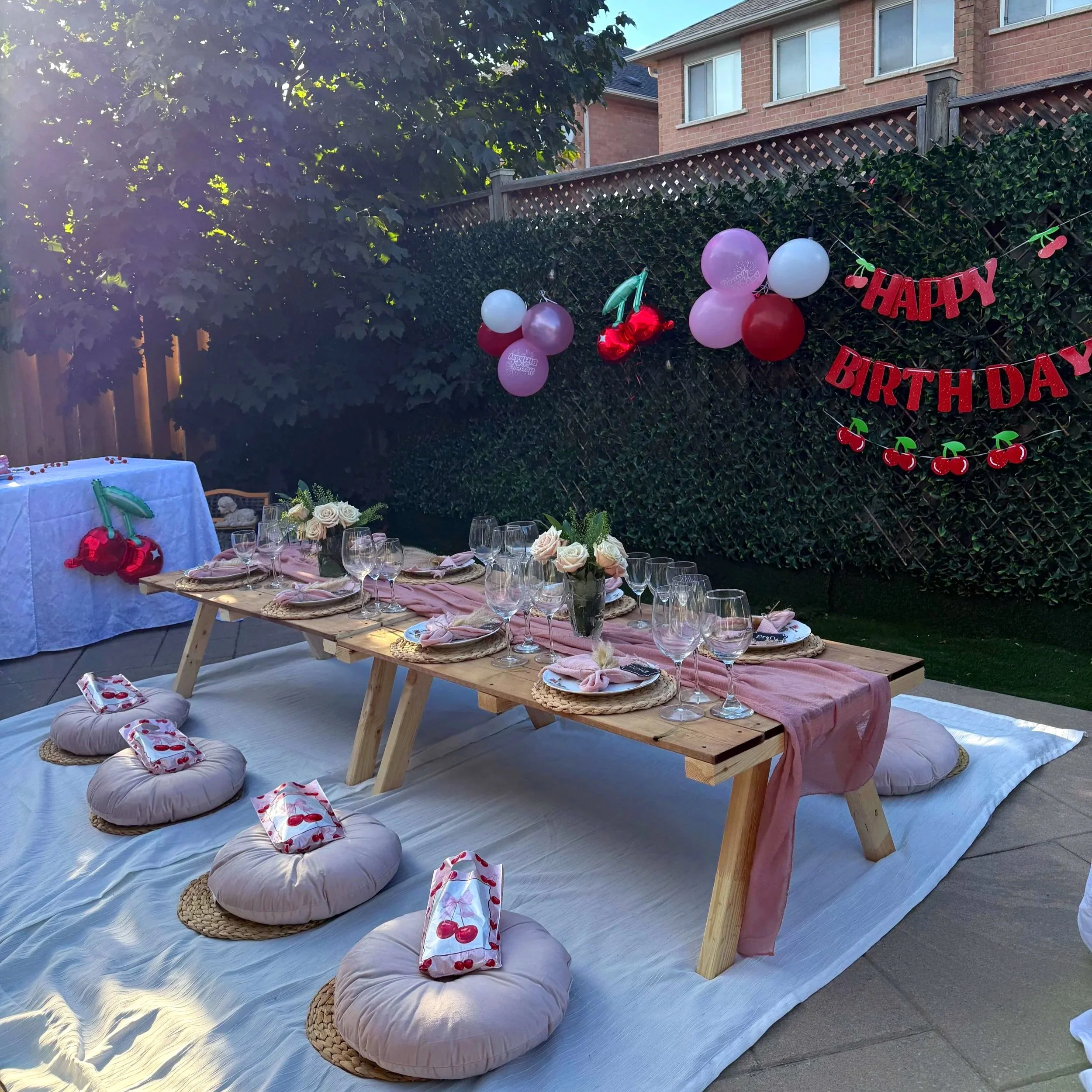 A backyard birthday celebration setup with a long wooden table covered in a pink table runner, surrounded by cushion seats on a white blanket. The table is set with wine glasses, plates, napkins, and floral centerpieces. A string of red and pink ball