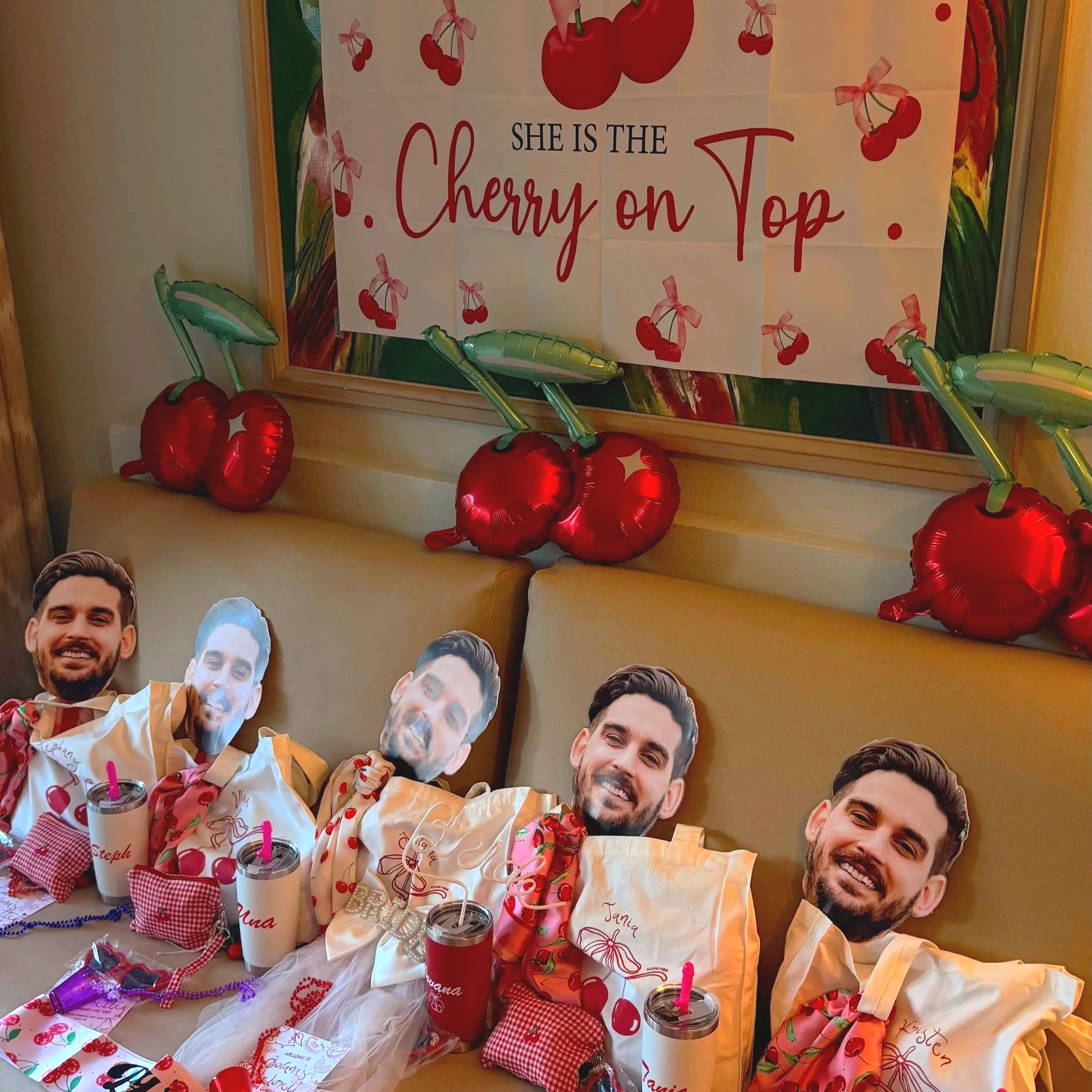 Colorful outdoor celebration with a large banner that reads, 'She is the cherry on top,' decorated with cherries and pink ribbons. The table below is filled with party favors, personalized drink cups with pink straws, small gift bags, and cutout phot