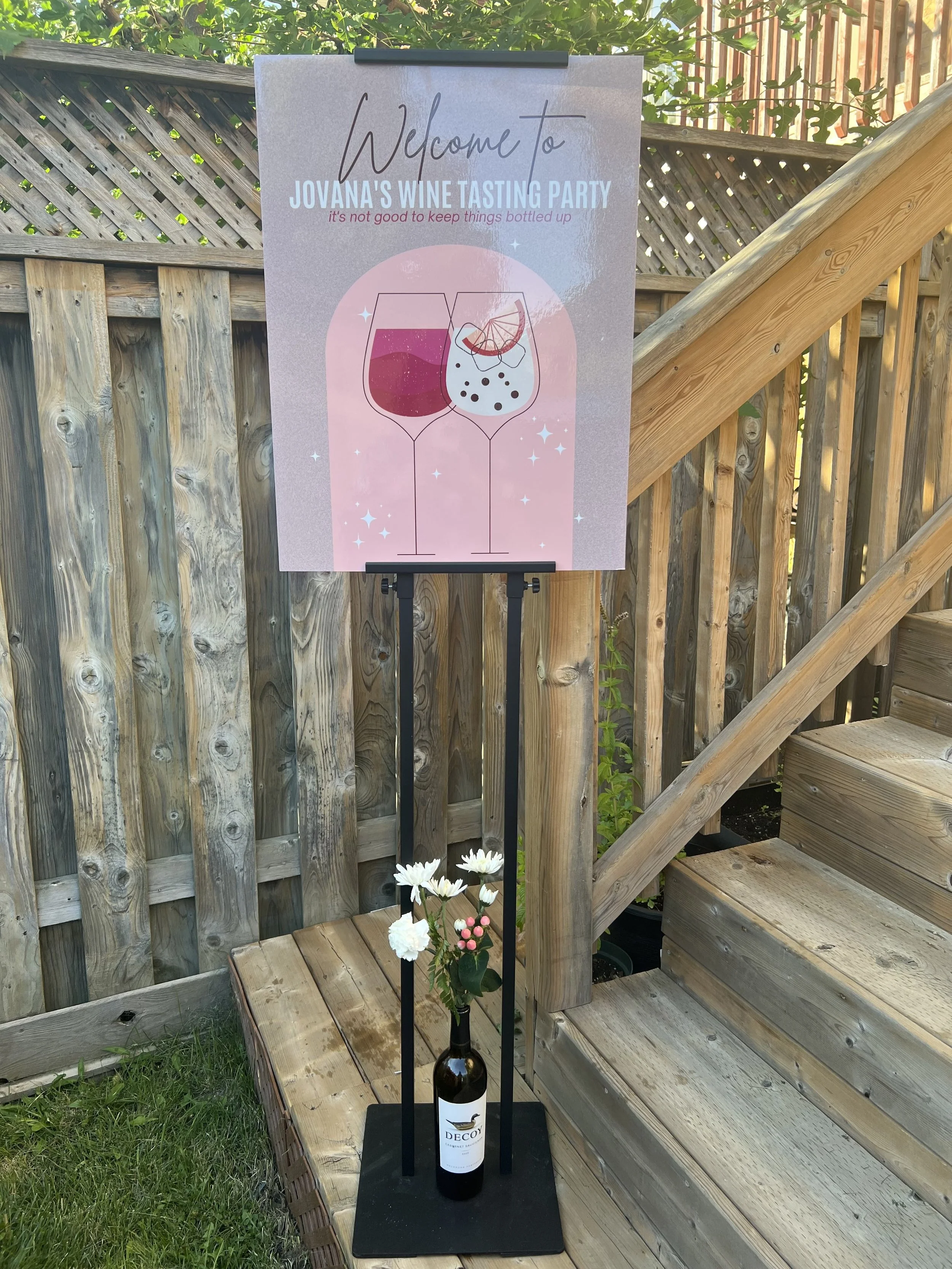 A welcome sign for Jovana's wine tasting party, featuring an illustration of two wine glasses, one red and one white, with a bottle of wine and white flowers at the base. The sign is outdoors near a wooden staircase and fence.