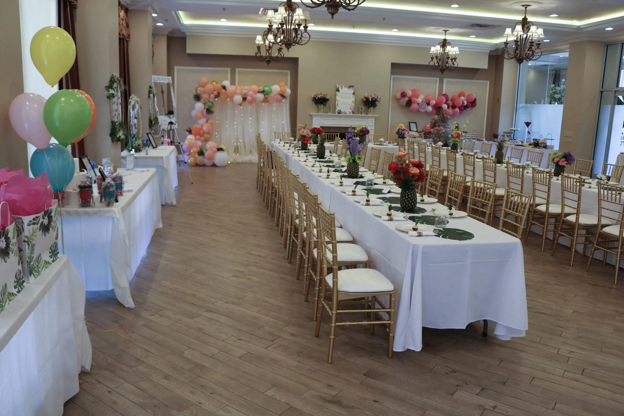 An event space decorated with pink and peach balloons, floral centerpieces, and elegant chandeliers, set up for a celebration or banquet.
