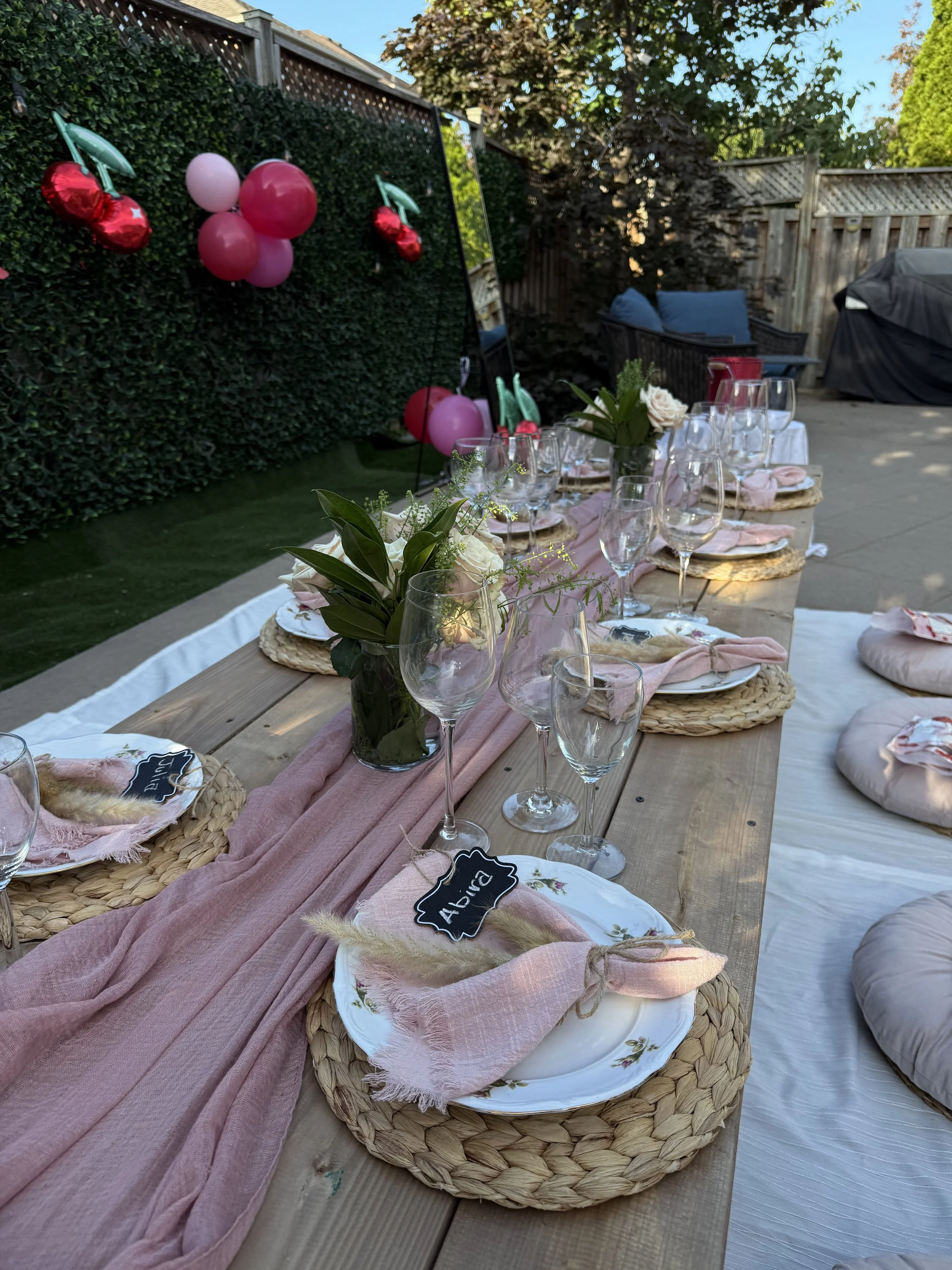 Outdoor table setting with pink table runner, glassware, floral arrangements, picnic plates, and cushioned seating, decorated for a celebration or gathering.