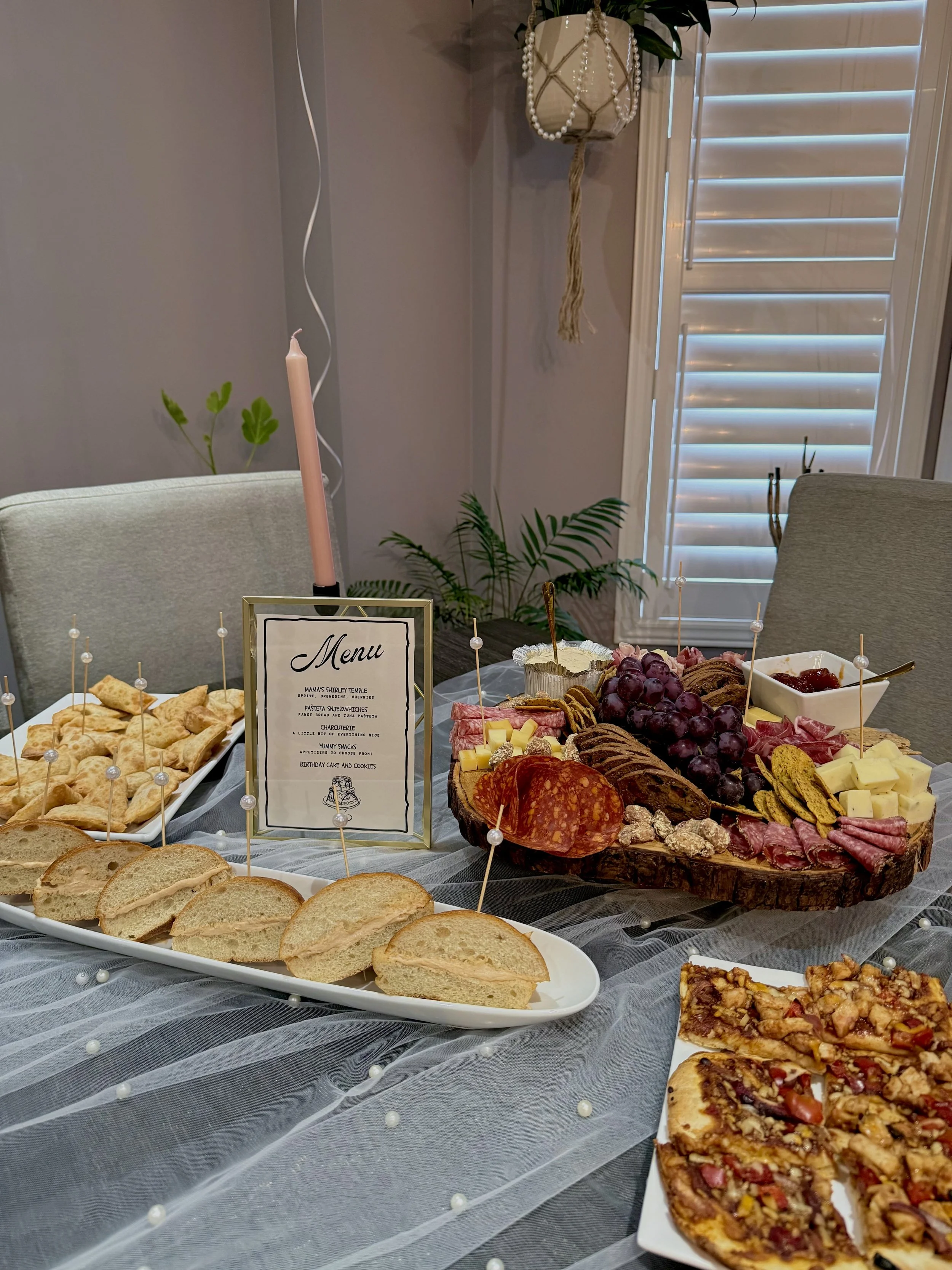 A buffet table with a variety of finger foods including sliced bread, crackers, a cheese platter with grapes and meats, and slices of pizza. The table is decorated with a sheer tablecloth and small pearl accents. In the background, there is a menu si