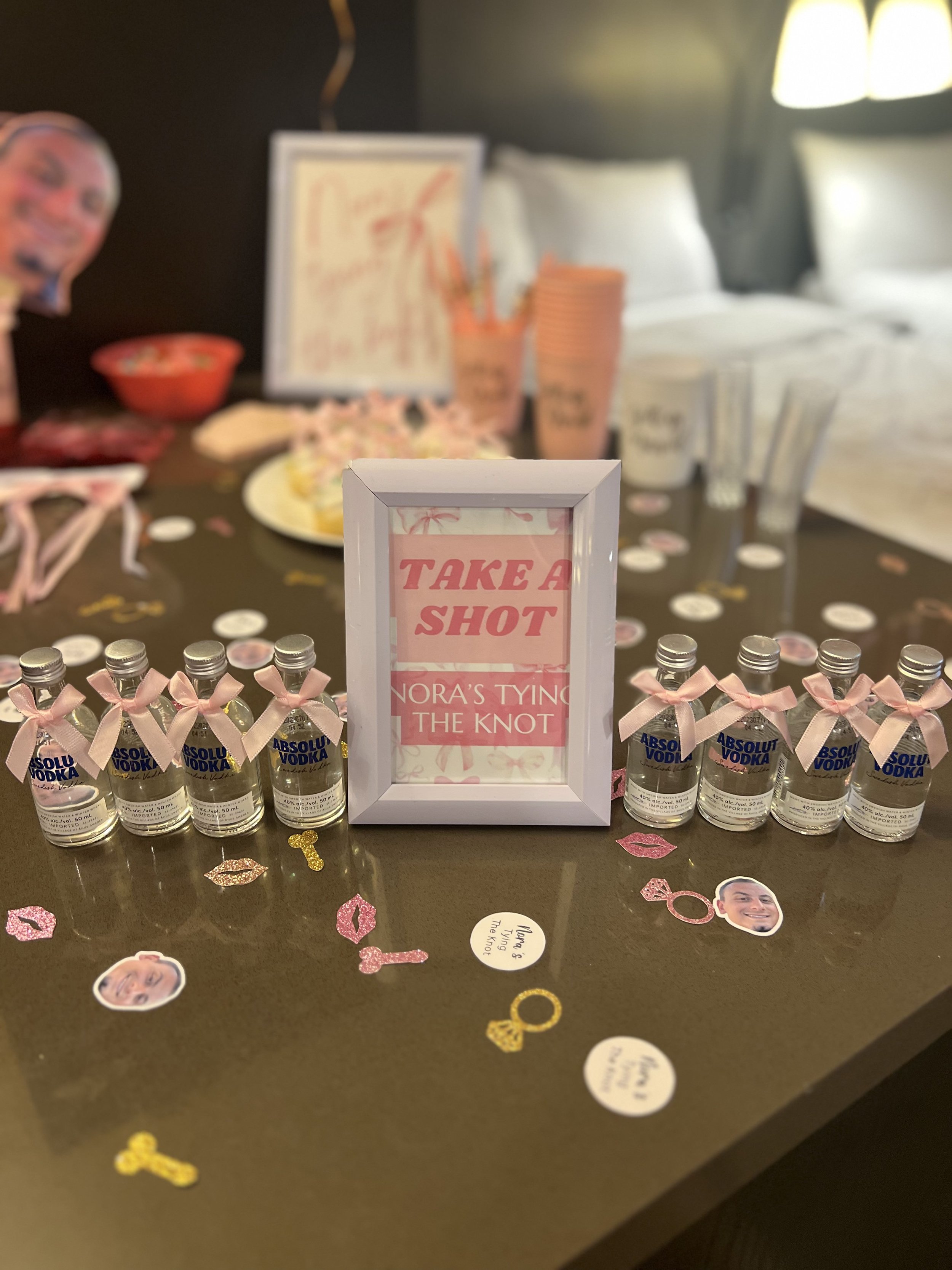 Table decorated for a celebration with a sign that says 'Take a Shot,' small bottles of Absolut vodka connected with pink ribbons, scattered paper cutouts of lips, and photos of a man and a woman.