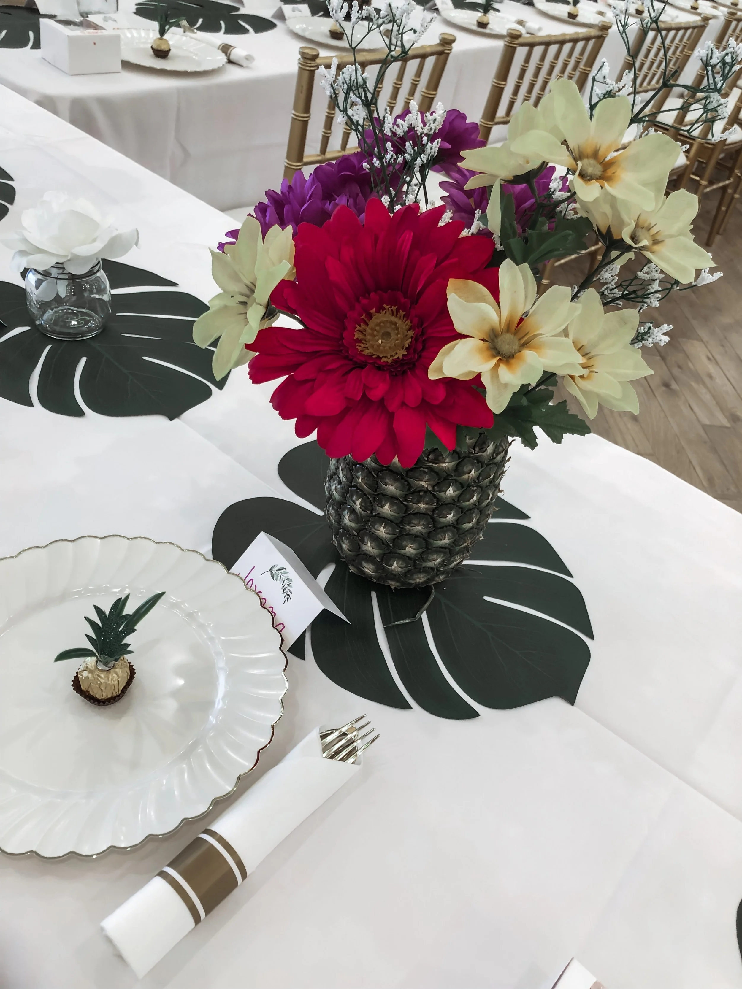 Decorative flower arrangement featuring a pineapple base with large red, purple, and cream flowers, accompanied by sprigs of white baby’s breath. The table setting includes white plates, a small glass vase with a white flower, gold-colored chairs, an
