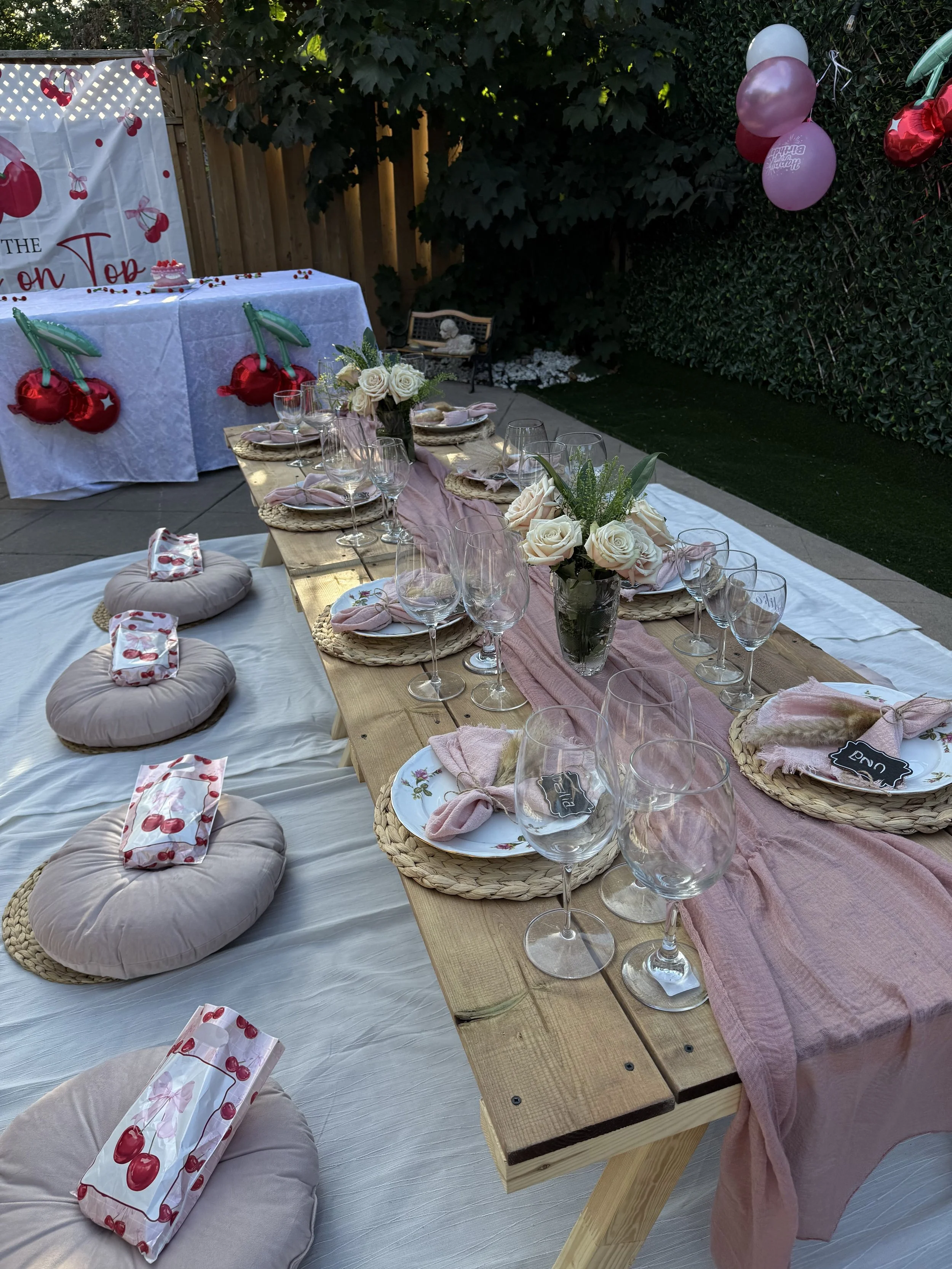 Outdoor birthday party setup with a long wooden table decorated with pink and white roses, glasses, plates, napkins, and small chalkboard name tags. Behind the table is a white tablecloth with cherry-themed decorations and a small birthday cake. To t