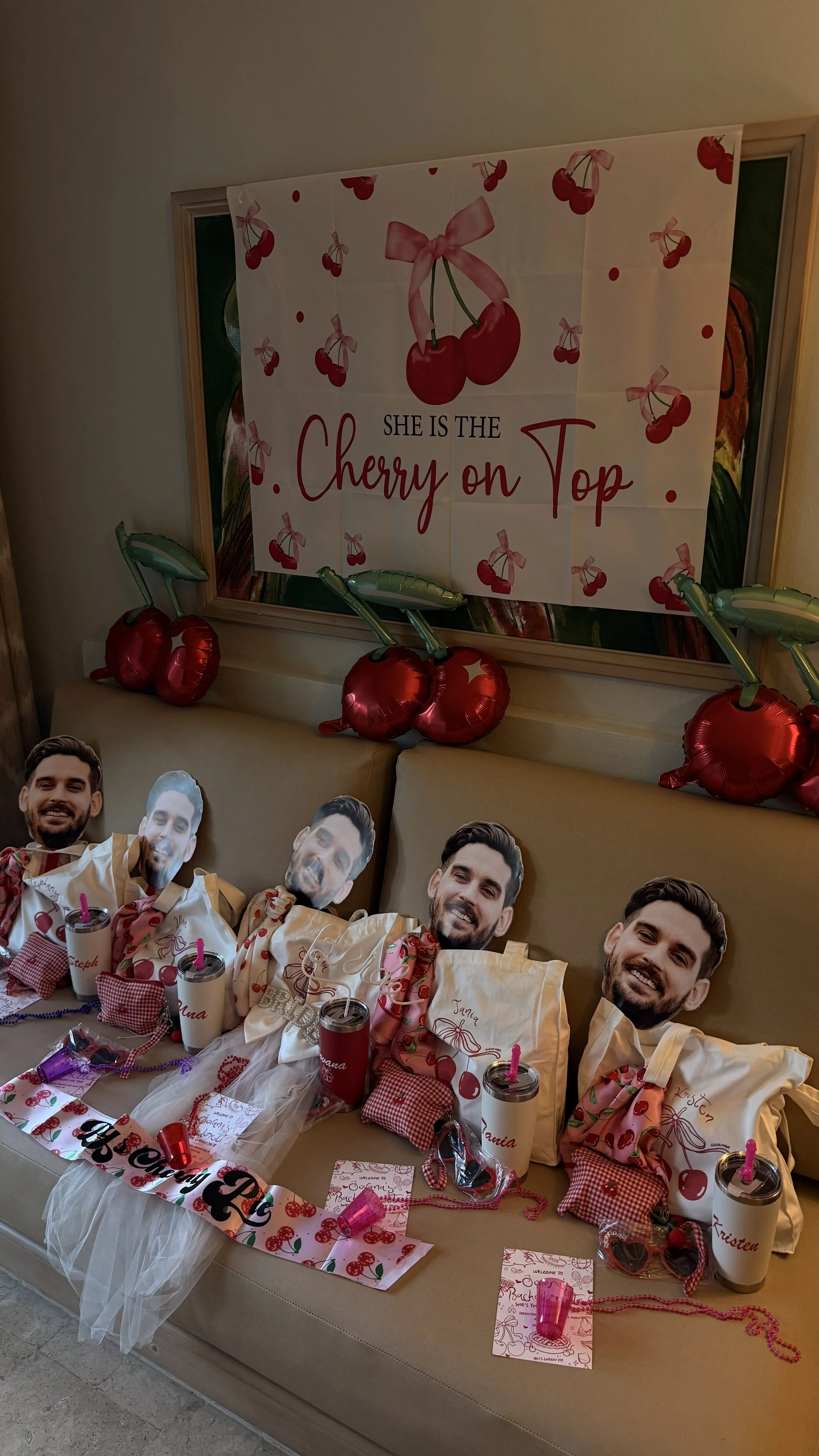 Decorated table with paper cutouts of a man's face, cherry-themed items, pink cups, and cherry balloons. There is a cherry-themed banner and a backdrop featuring cherries and the text 'She is the cherry on Top' with cherry illustrations.