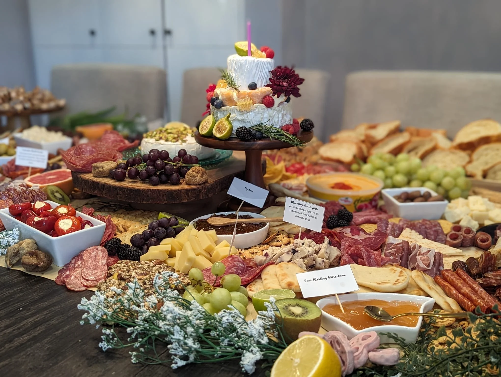 A buffet table with a multi-tiered cake decorated with fruits and flowers, surrounded by various cheeses, meats, fruits, and condiments, with small signage labels.