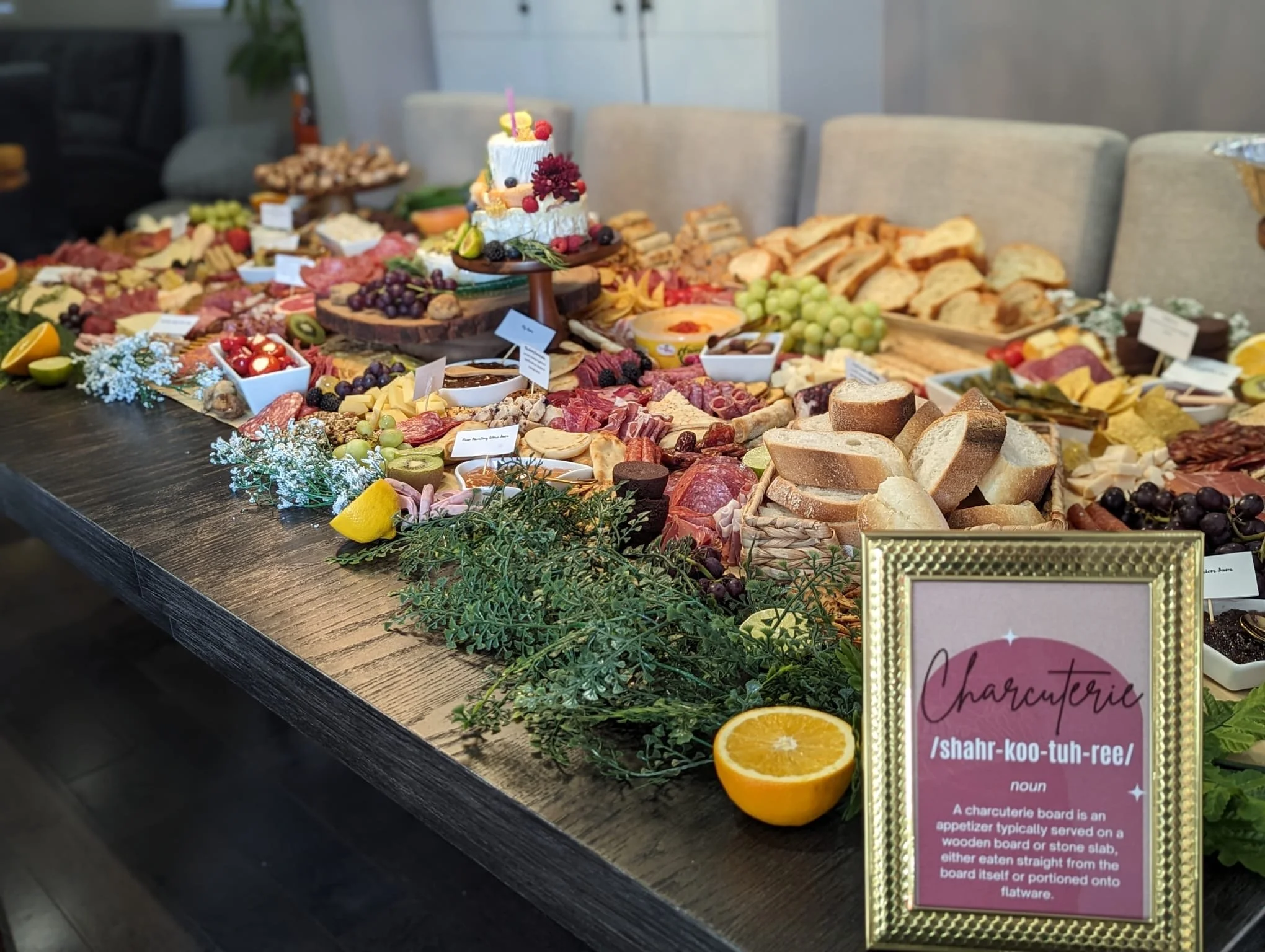 A large wooden table filled with various cheeses, sliced meats, fruits, bread, and small bowls of spreads, arranged for a charcuterie board display. A small decorative cake and greenery are in the center, with a pink framed informational sign in the 