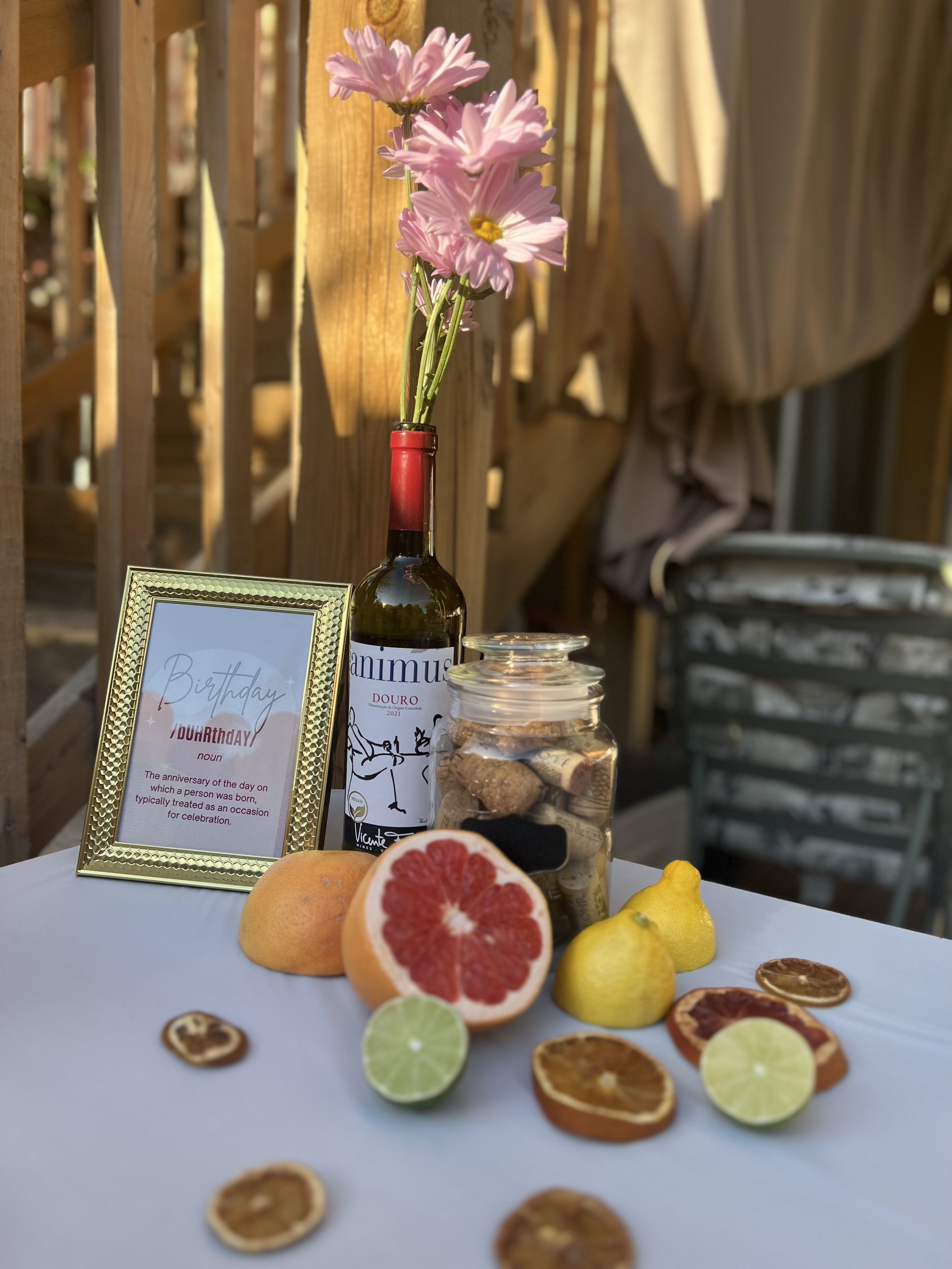 A decorative table setting featuring a bottle with pink flowers, a gold picture frame with a birthday message, slices of citrus fruits, a grapefruit, lemons, limes, and dried fruit slices, all set against a wooden background.