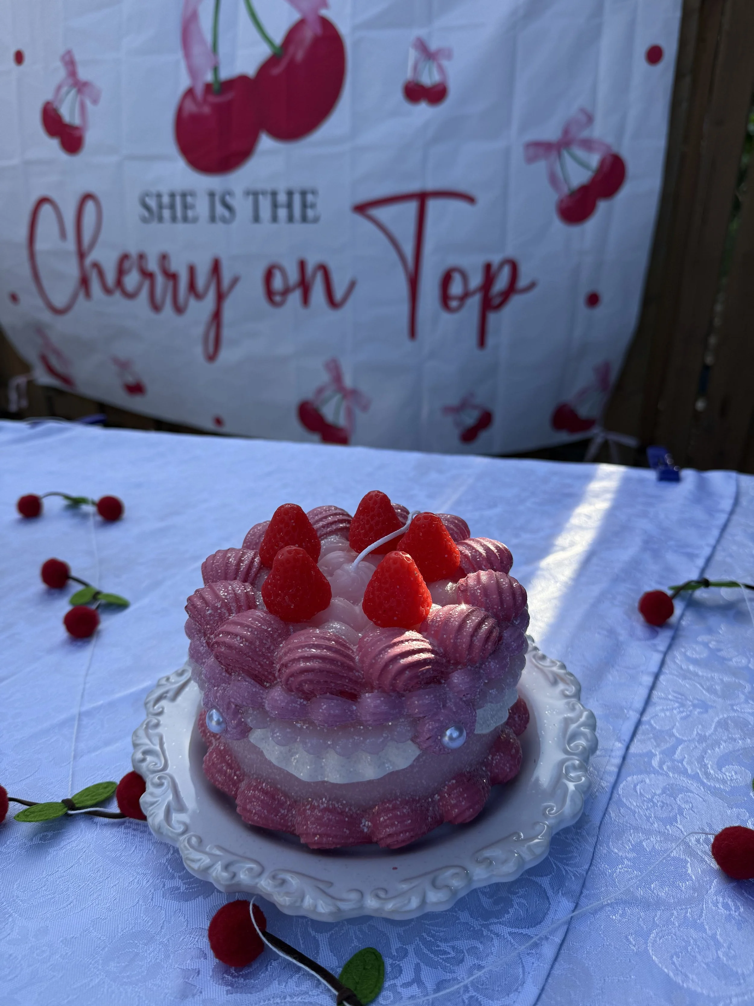 A pink and white cake topped with fresh strawberries and pink cookies, placed on a white ornate plate on a white tablecloth with cherries and green leaves decorating it, with a cherry-themed sign in the background.