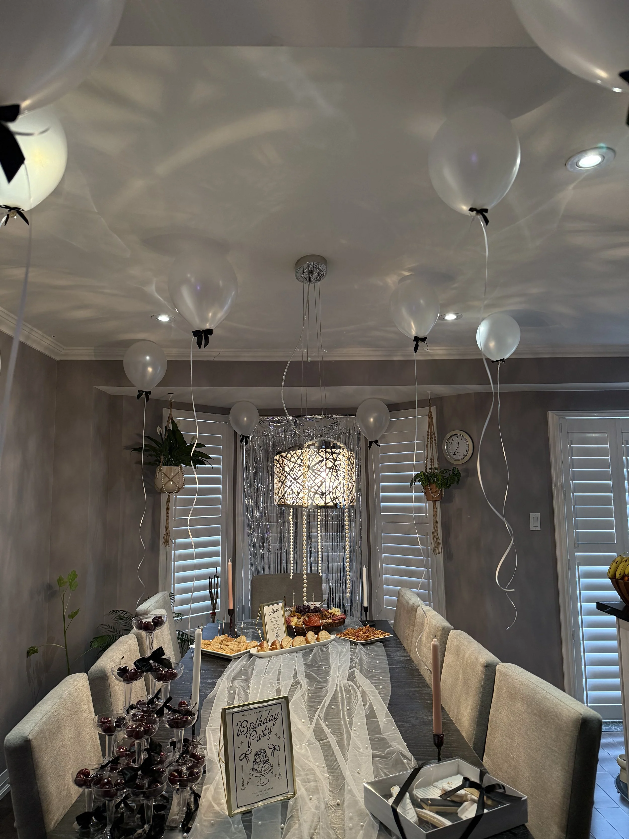 A dining room decorated for a birthday party with balloons hanging from the ceiling, a table covered with a white tablecloth, and various desserts and snacks. There are chairs around the table, and a sign on the table says 'Birthday Party'.