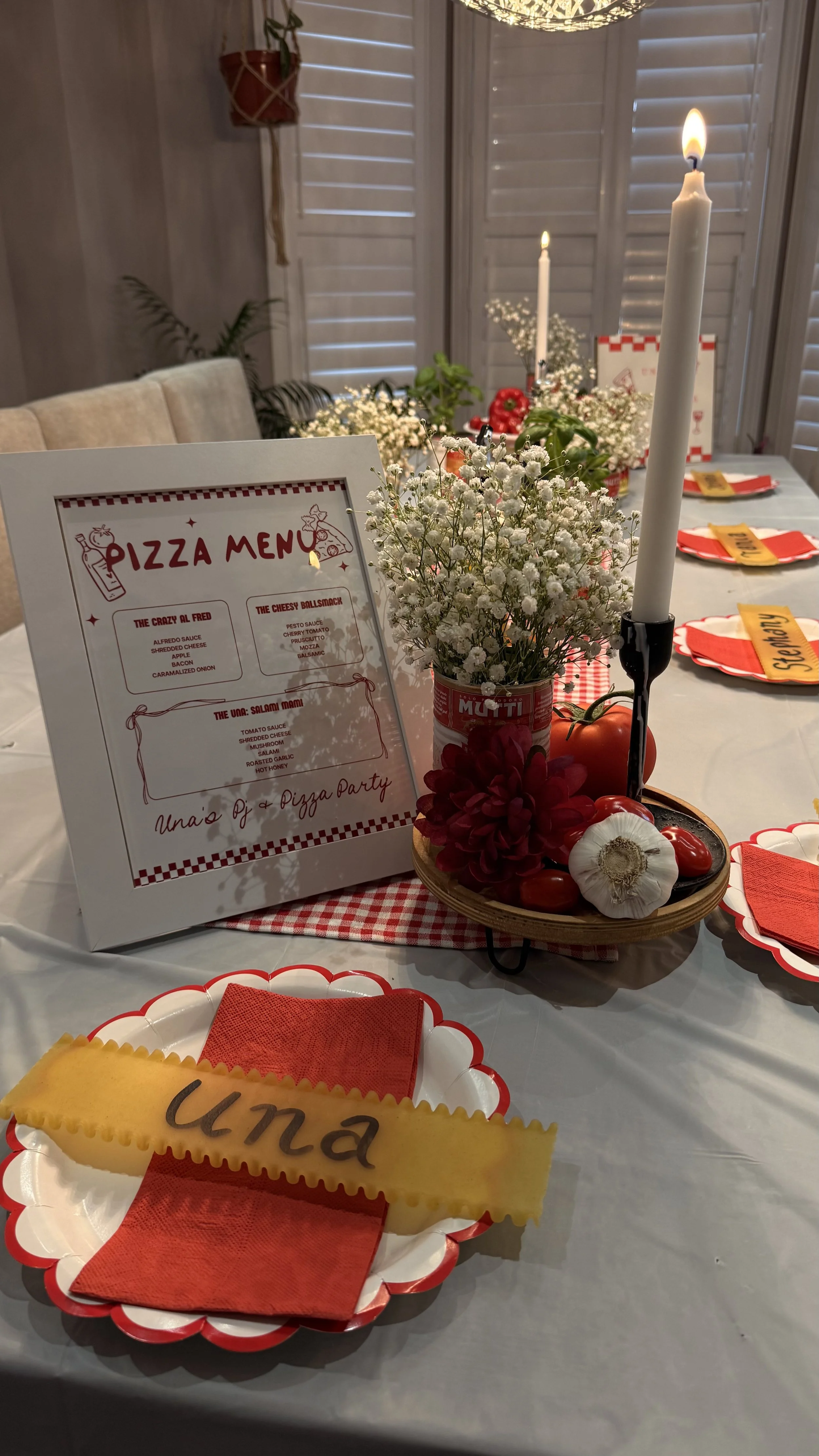 A decorated dining table with a pizza menu, flowers, candles, and a plate with a pizza slice and the name 'Ula' written on it, in a cozy room with wooden shutters and hanging plant.