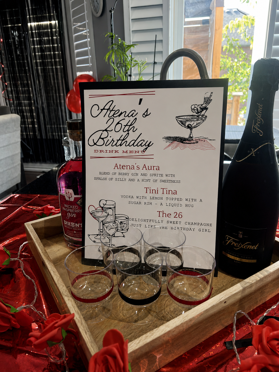 A birthday drink menu for Atena's 26th birthday, featuring three cocktails: Atena's Aura, Tini Tina, and The 26, with illustrations of drinks on the menu. The menu is placed inside a wooden tray with glasses, a bottle of Wicked Raspberry Gin, and a large bottle of Freixenet sparkling wine. The background shows a window, some greenery, and decorative red roses and string lights around the tray.
