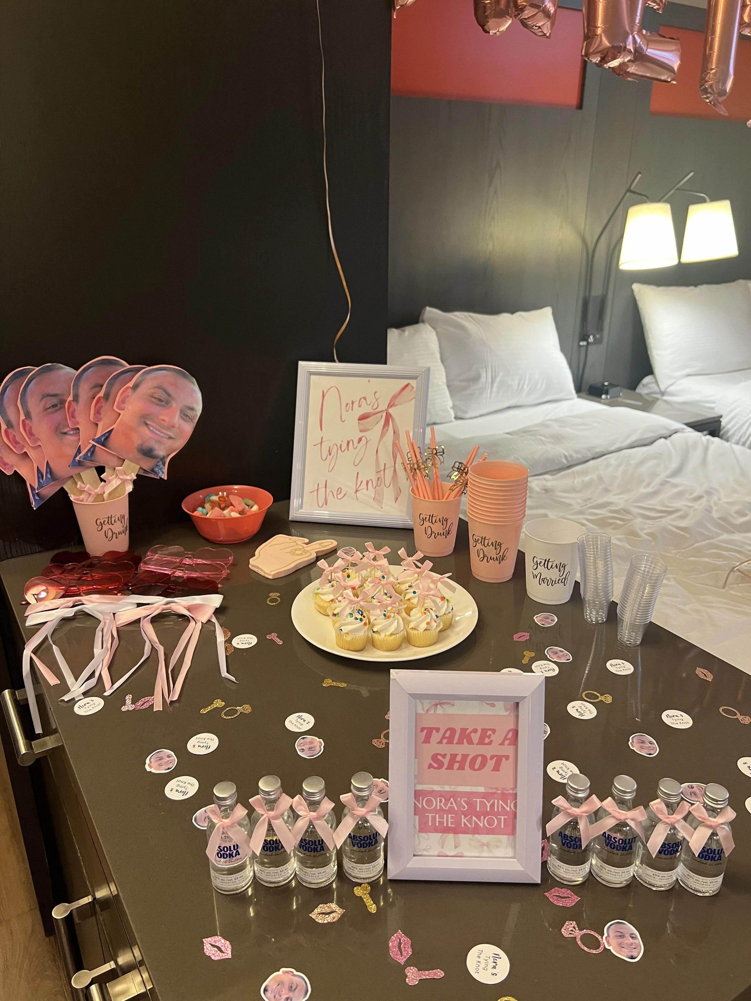 Party setup with cupcakes, heart-shaped cookies, pink and white ribbons, and bottles of vodka. Decorations include photo cutouts of a man with humorous text, pink lips cutouts, and a sign that reads 'Take a Shot'. There are also balloons and a bed wi