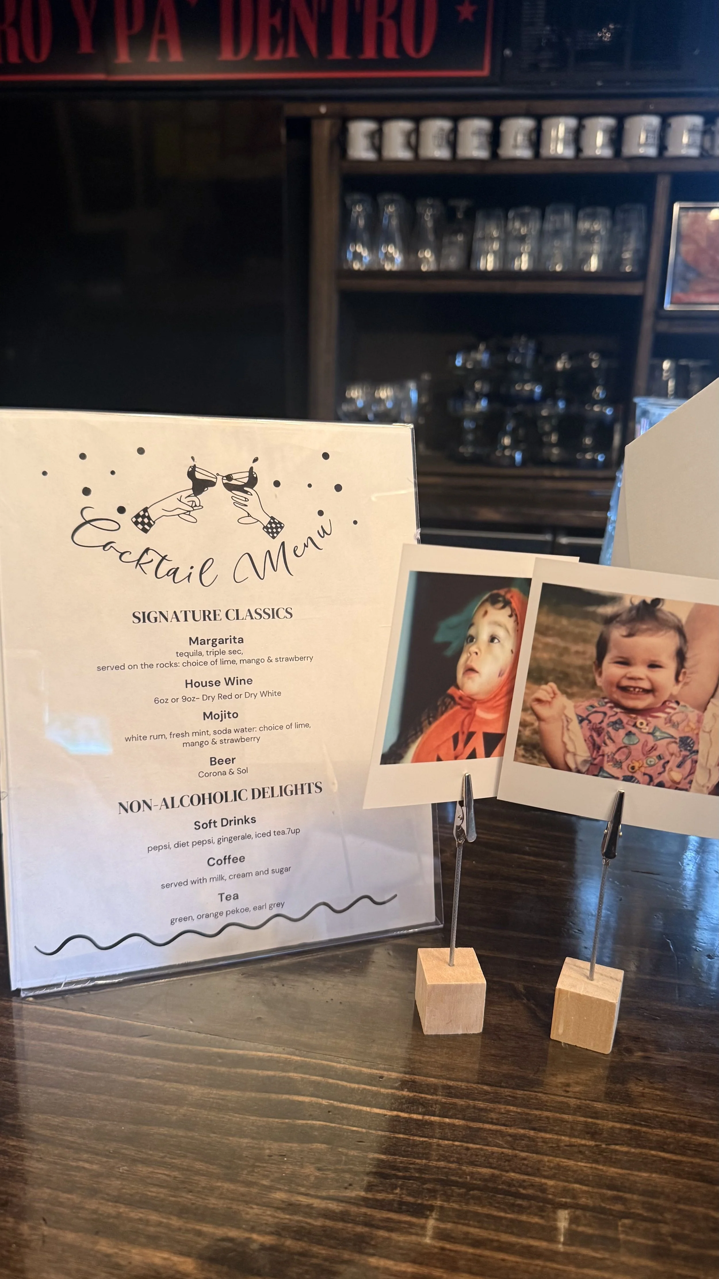 A cocktail menu with illustrations of glasses clinking, listing signature drinks such as Margarita, House Wine, and Mojito, alongside photographs of children and a wooden table.