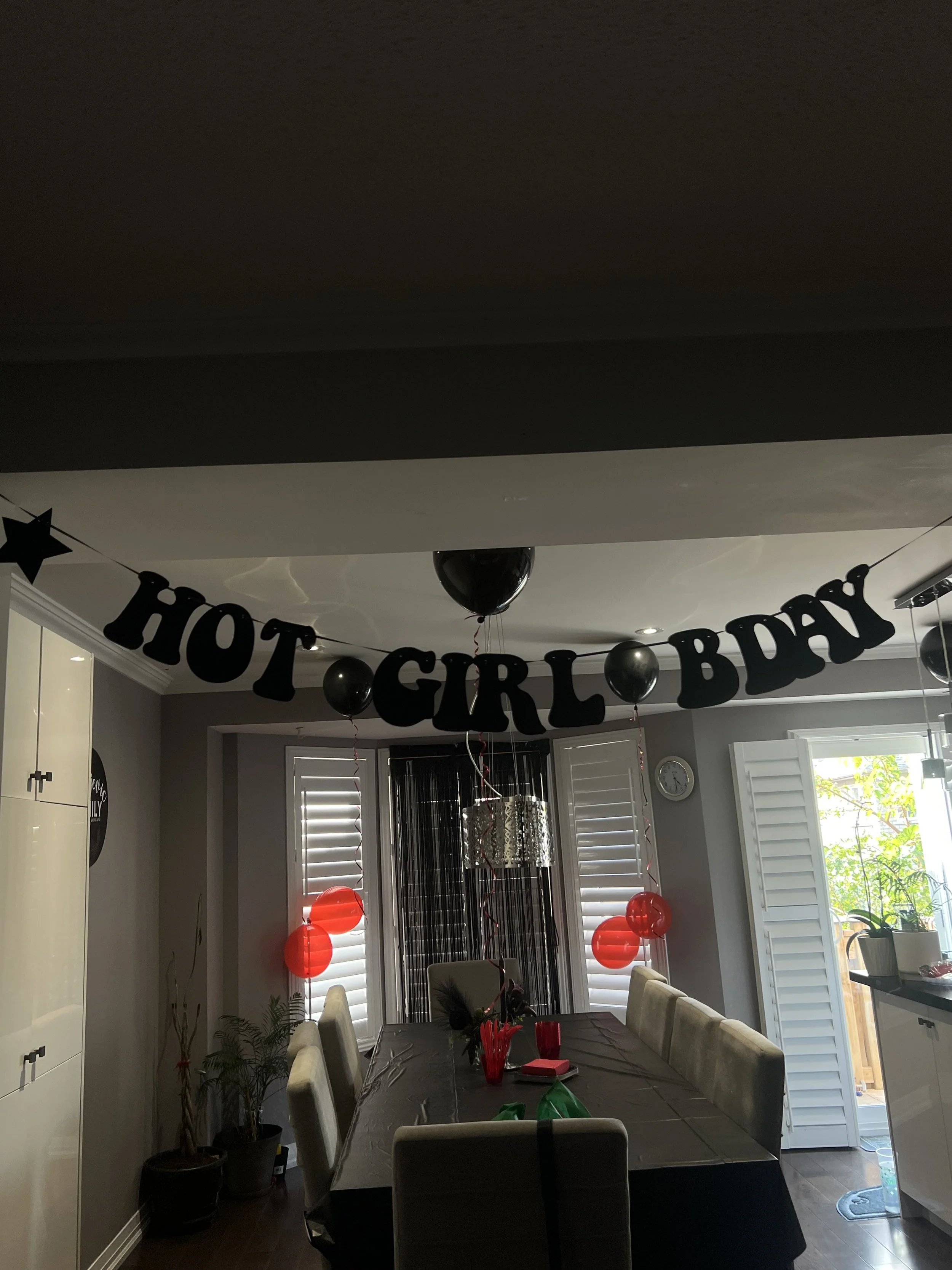 A dining room decorated for a birthday celebration with a 'Happy Birthday' banner, red and black balloons, and a table set with party supplies.