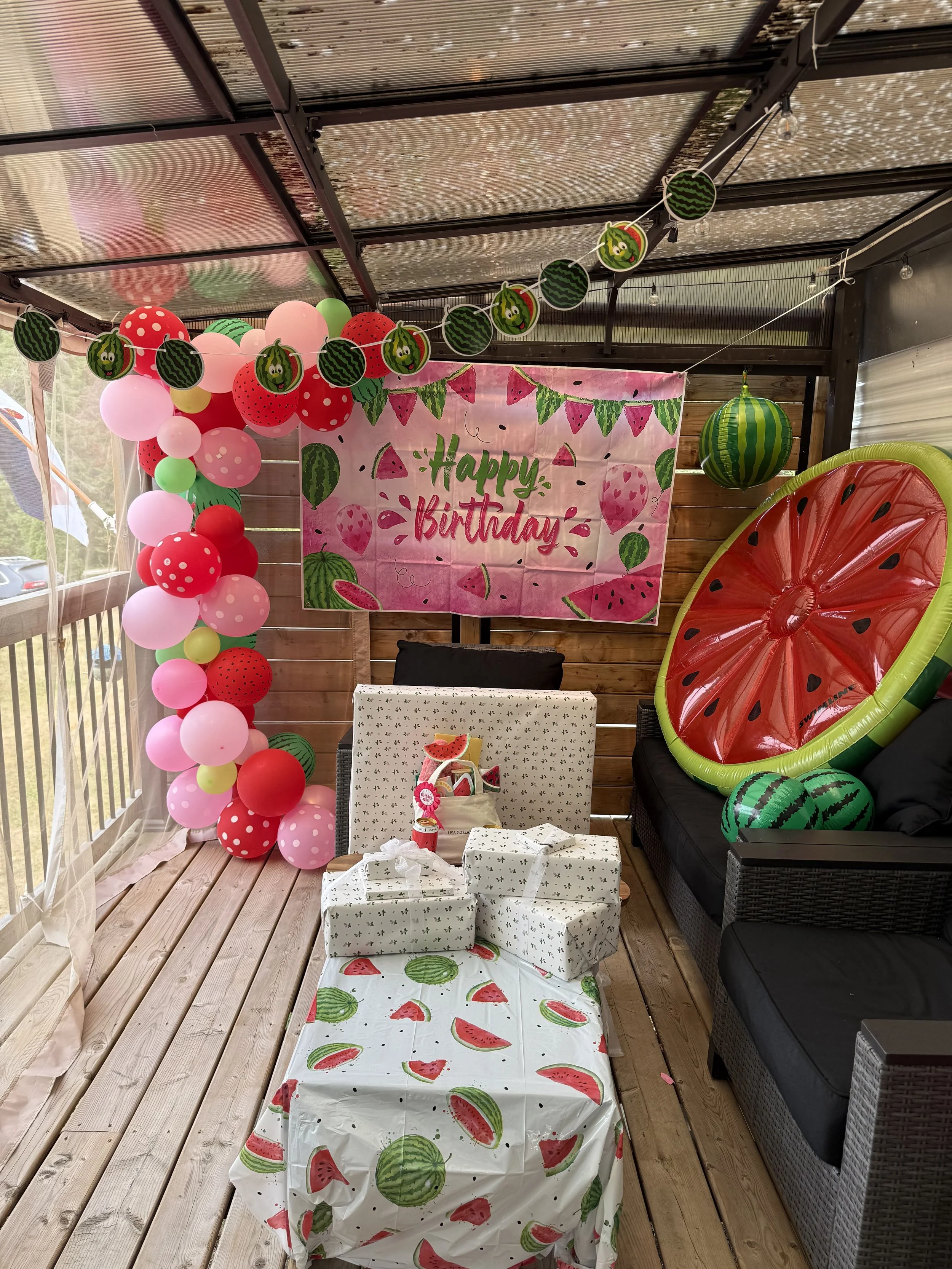 Decorated porch with watermelon themed decorations for a birthday party, including pink, red, and green balloons, a watermelon inflatable, a watermelon-themed tablecloth, a large watermelon hanging decoration, and a pink banner that says 'Happy Birth