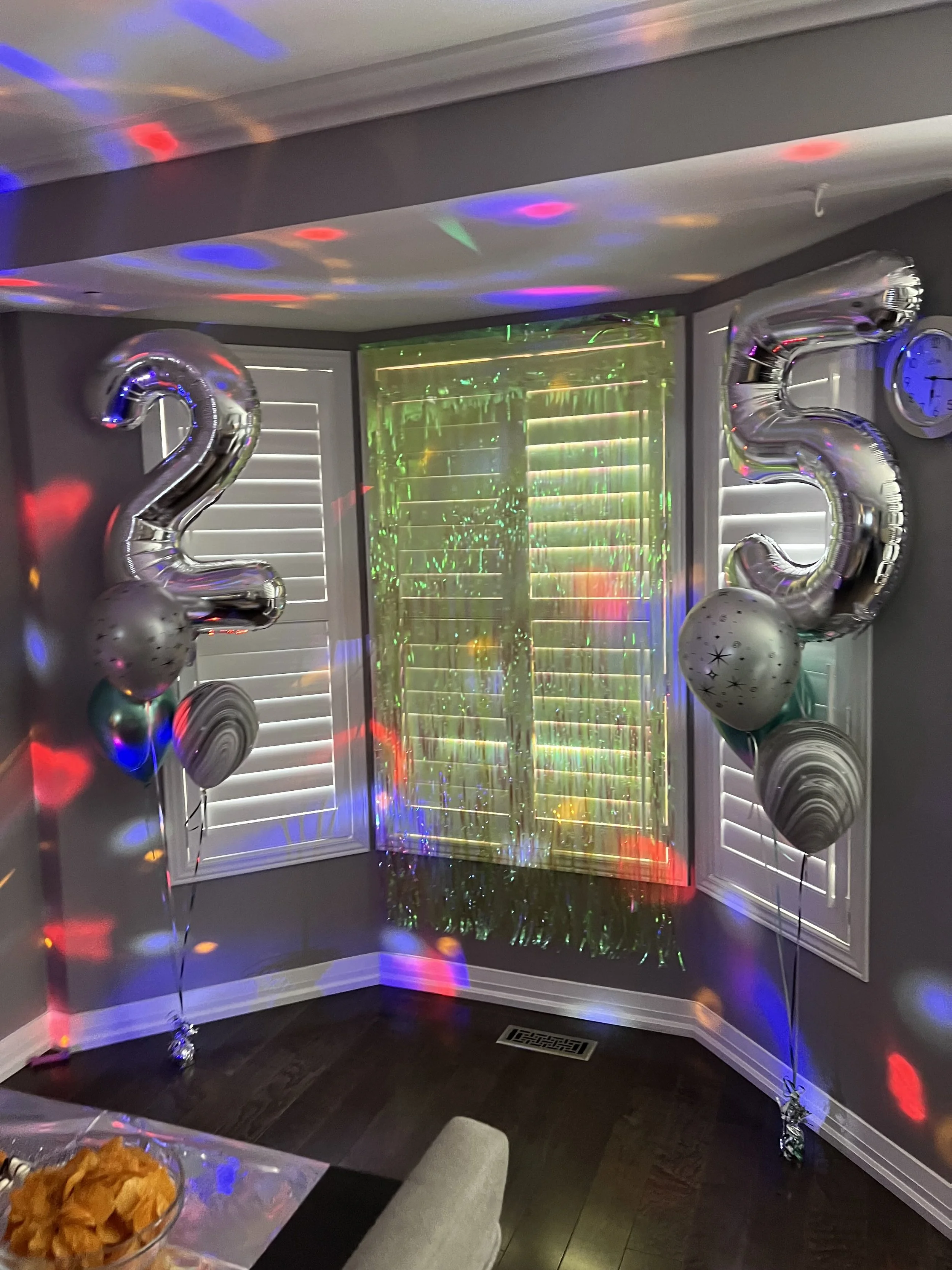 Living room decorated for New Year's celebration with large silver balloons forming the numbers 2023, balloons with star and swirl patterns, colorful disco lights projecting patches of red, blue, green, and purple, a table with snacks in the foregrou