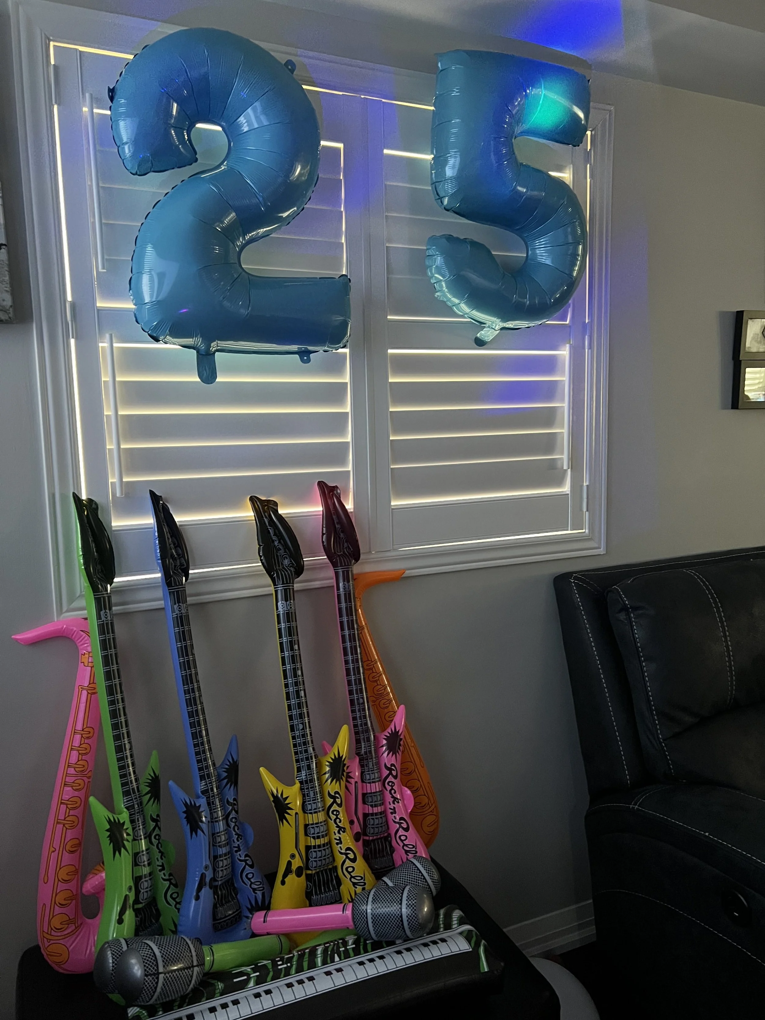 Colorful inflatable musical instruments including two numbers '2' and '5' balloons, several music-themed balloons shaped like microphones and instruments, and a row of toy guitars leaning against a black sofa in a room with white blinds and a wall mi