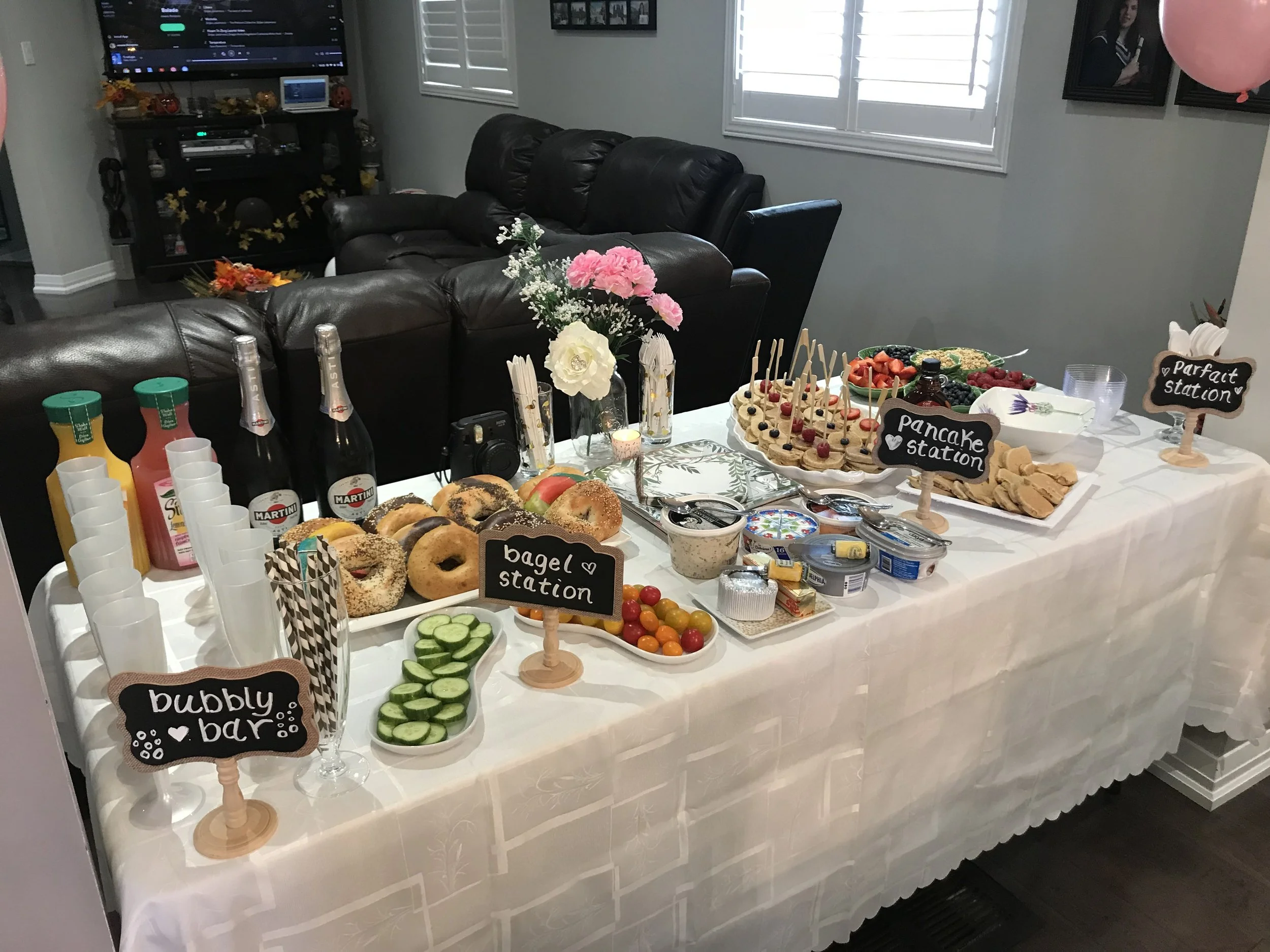 A table set up for a celebration with various food and drinks including donuts, fruit, and condiments, decorated with pink flowers and balloons.