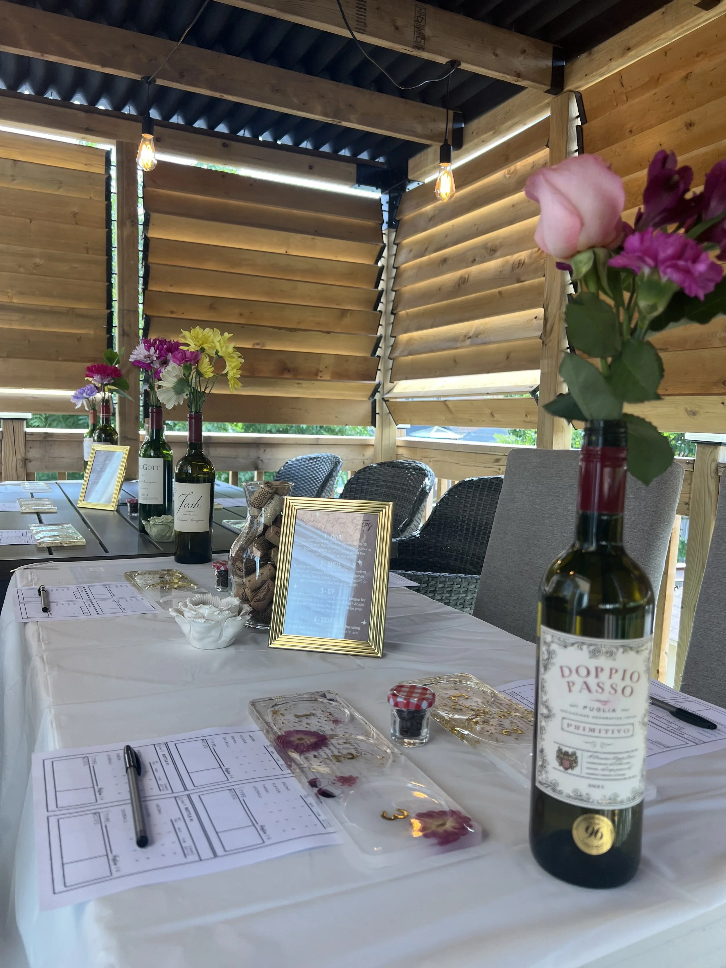 A table set for a tasting or event with flower arrangements in wine bottles, framed signs, papers, pens, and small plastic containers, inside a wooden structure with horizontal wood panel walls and decorative lighting.