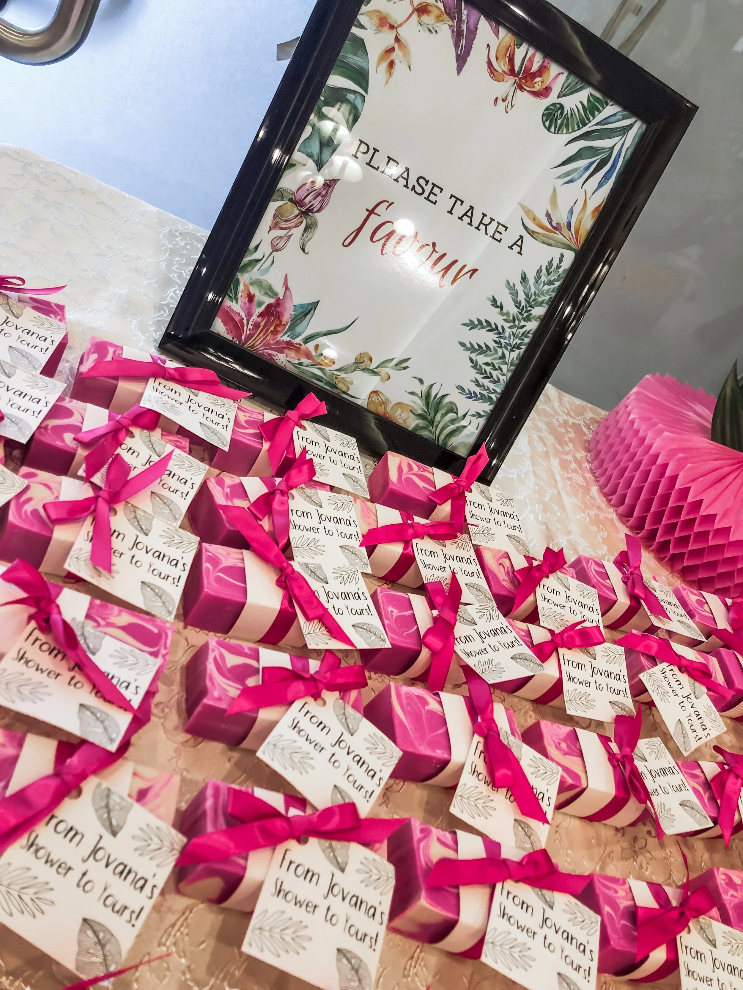 A display of pink gift boxes with white tags that read 'From Jovana's Shower to Yours!' tied with pink ribbons, set on a table with a pink ruffled decorative piece. Behind the boxes, there is a framed sign that says 'Please take a favor' with floral 