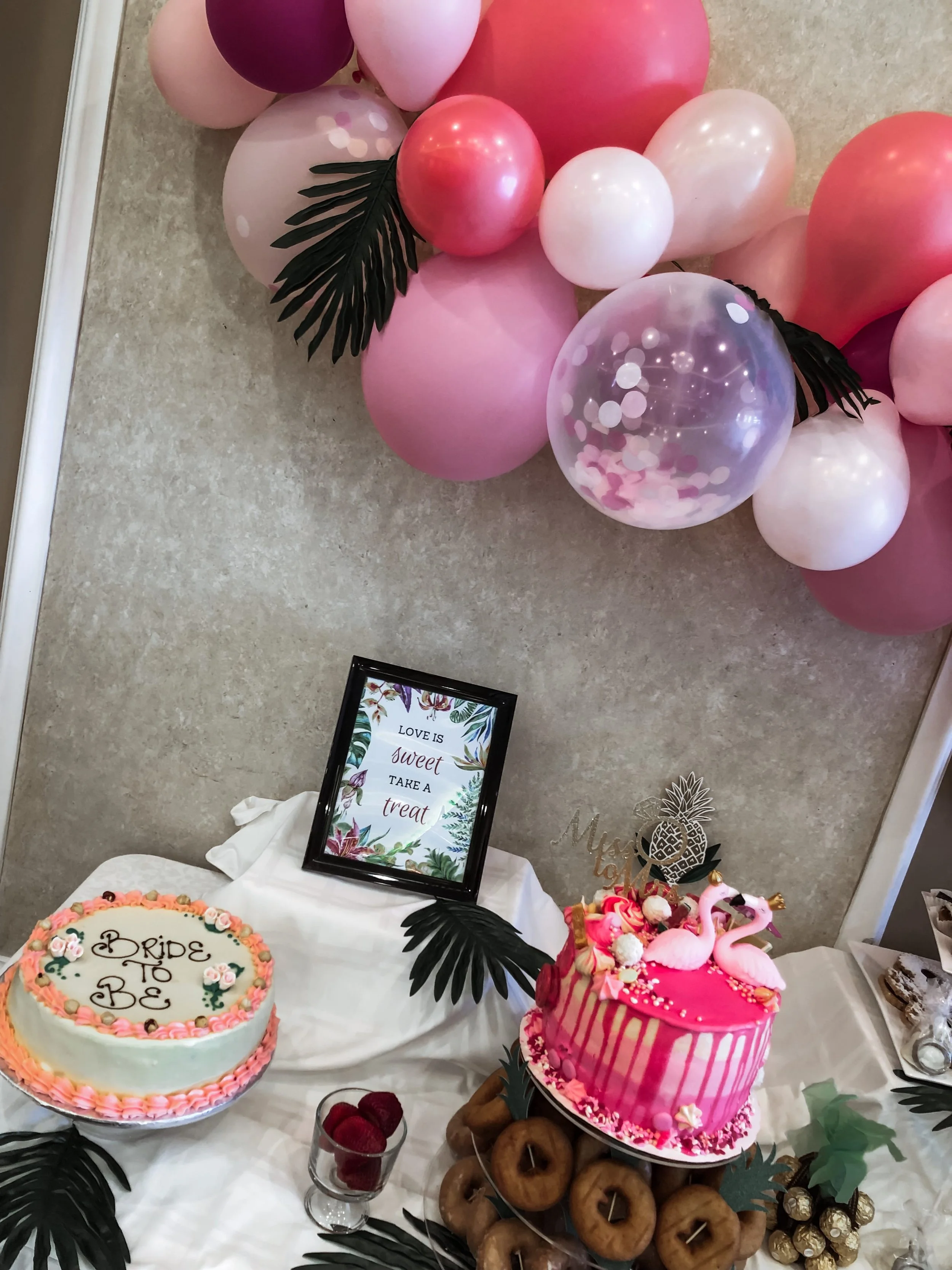 Decorative dessert table with pink and white balloons, a cake with the words 'Bride to Be', pink flamingo decoration, and a framed sign that reads 'Love is sweet, take a treat' at a celebration event.
