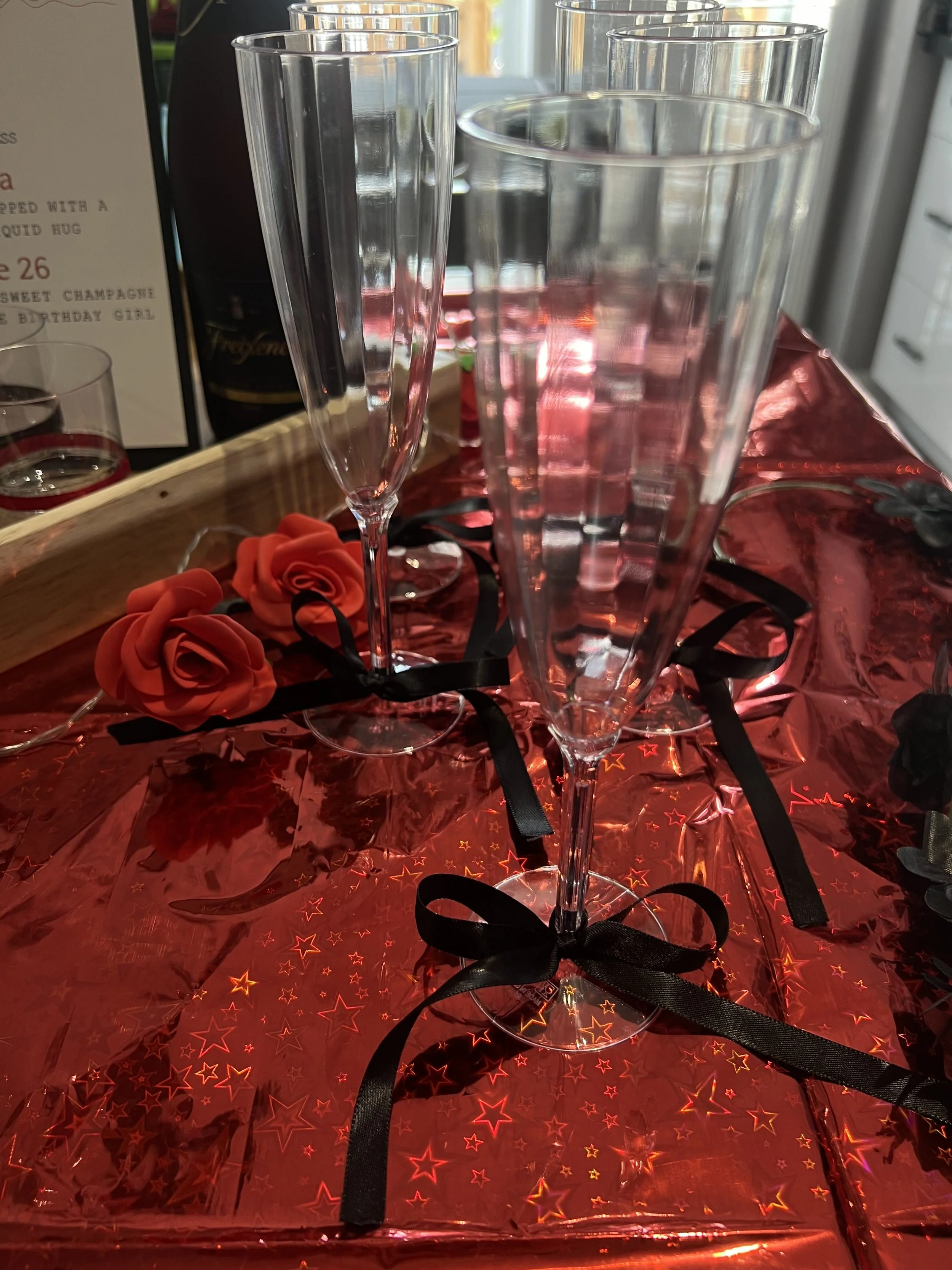 Multiple empty clear drinking glasses on a red star-patterned tablecloth, with some decorated red artificial roses and black ribbon decorations, alongside a wooden tray holding a menu and a bottle of champagne in the background.
